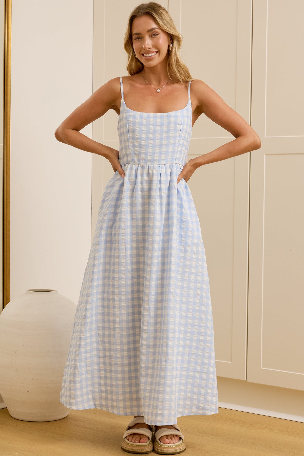 A woman modeling a light blue and white gingham maxi dress with spaghetti straps in a bright room with cream-colored wardrobes and a large mirror showing her reflection.