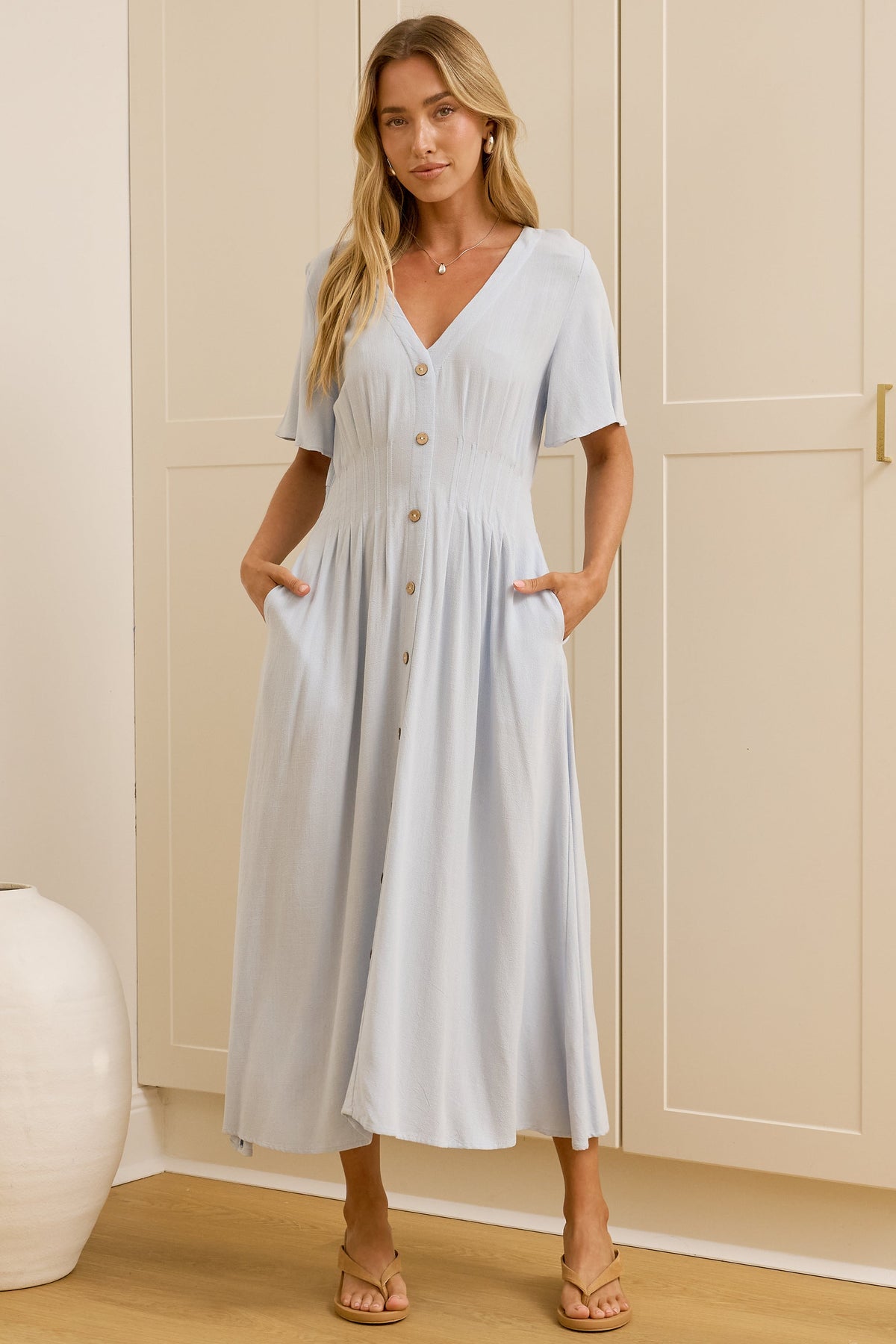 A woman modeling a light blue midi dress with short sleeves, a V-neckline, and front buttons, standing in a room with cream-colored cabinets and a mirror showing her side profile.