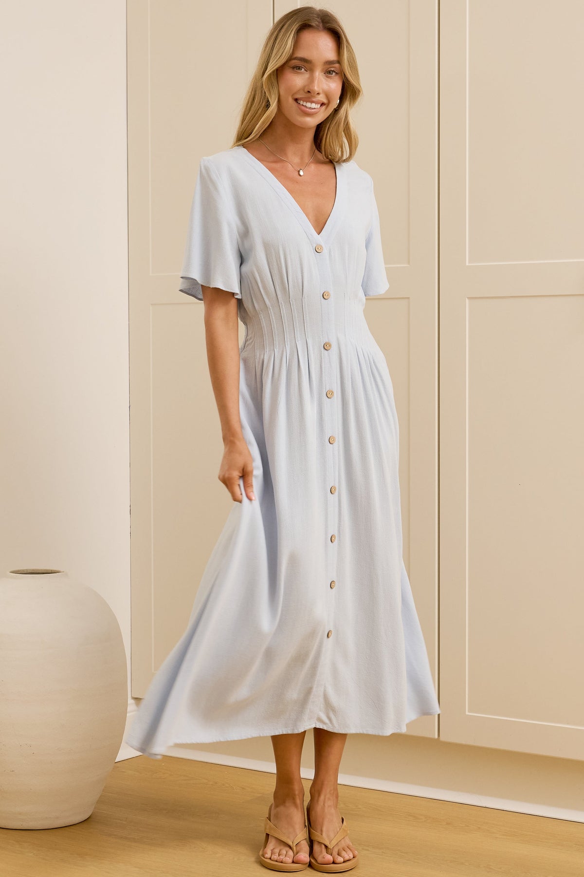 A woman wearing a light blue midi dress with short flutter sleeves and a button-front V-neck stands in a bright room with cream-colored cabinets, with her reflection visible in a mirror on the left.