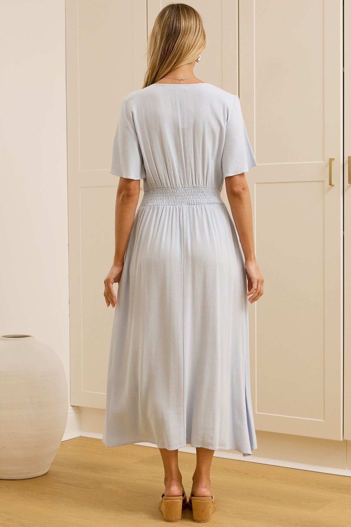 A woman stands with her back to the camera wearing a light blue midi dress in a minimalist room with cream-colored wardrobes and a large mirror showing her side profile.