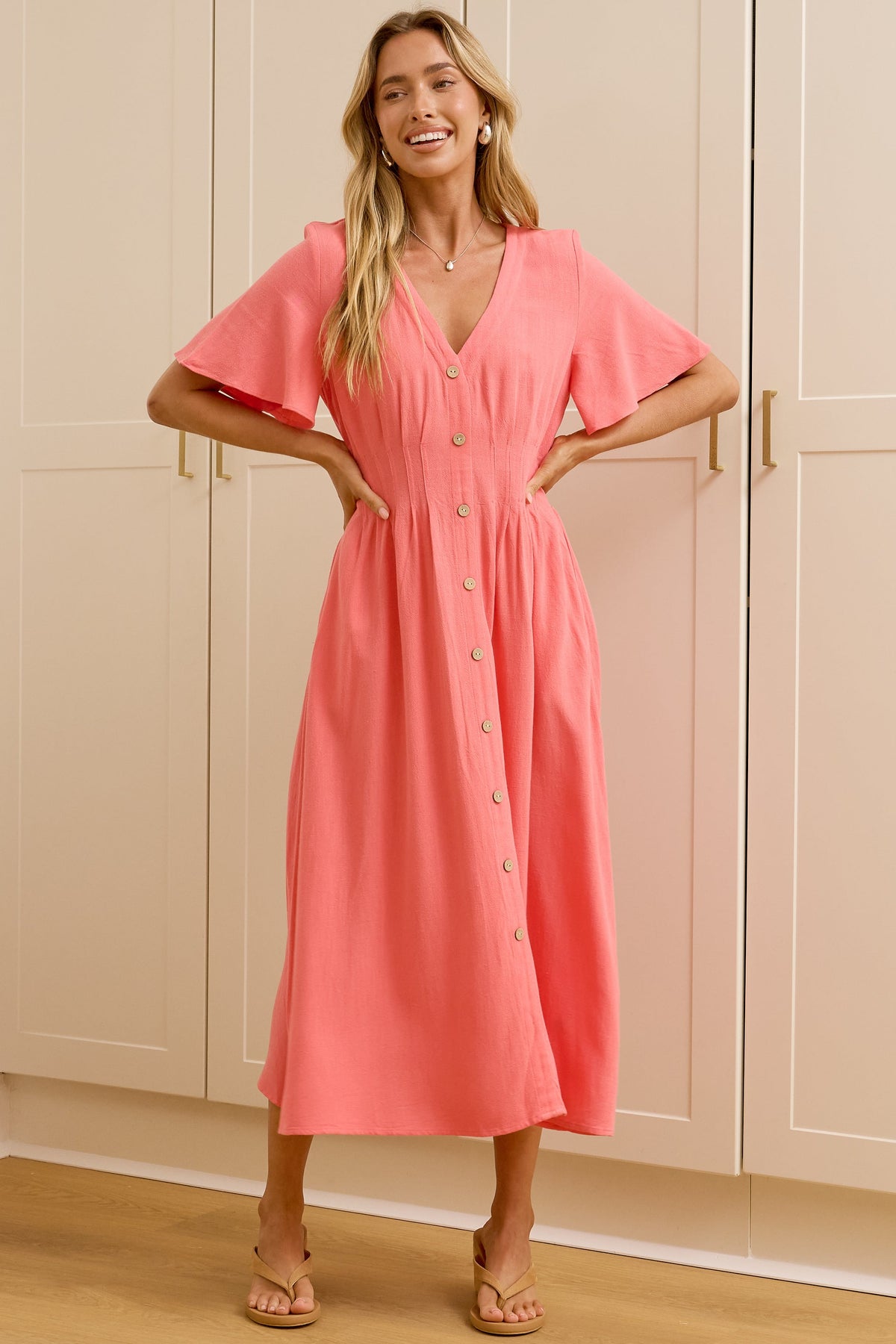 A woman stands in a bright room wearing a coral pink midi wrap dress with short flutter sleeves and a button-front waist. She is posed in front of white built-in wardrobes, with a mirror on the left reflecting her side profile.