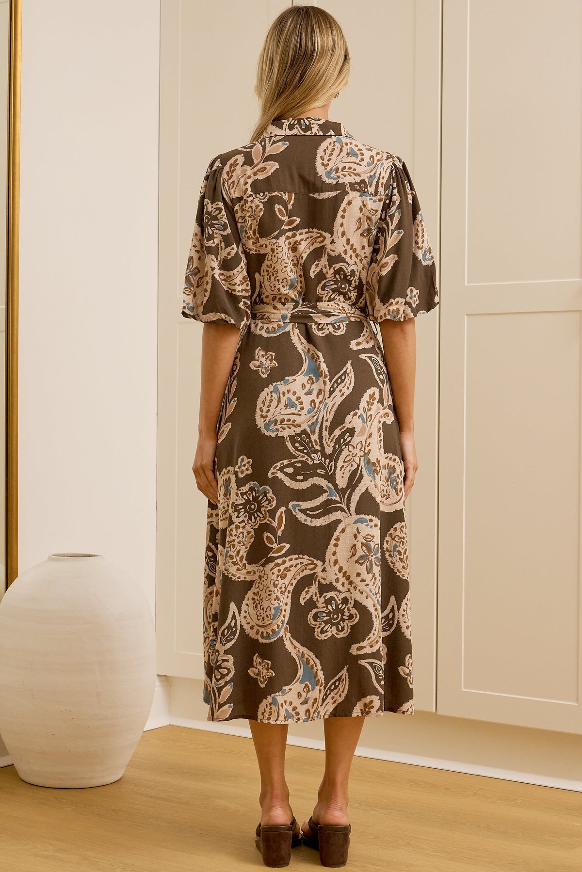 A woman stands with her back to the camera wearing a brown short-sleeved midi dress with a beige and blue paisley pattern, standing in front of white built-in wardrobes. A large floor-to-ceiling mirror on the left reflects her side profile.