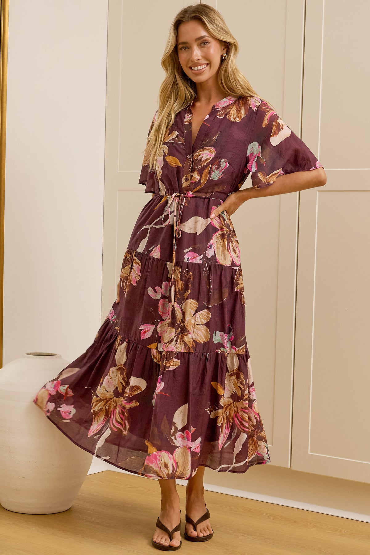 A woman models a tiered, short-sleeved floral maxi dress in a dusty purple shade with pink and yellow floral patterns. She is standing in a bright room with cream-colored paneled walls, a large floor-to-ceiling mirror showing her side profile, and a white ceramic vase on a light wood floor.