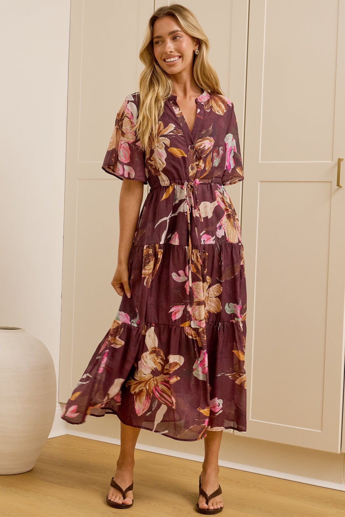 A woman models a purple floral midi wrap dress in a bright room with cream-colored wardrobes and a large mirror showing her profile. The dress features a purple base with pink, orange, and yellow floral patterns.