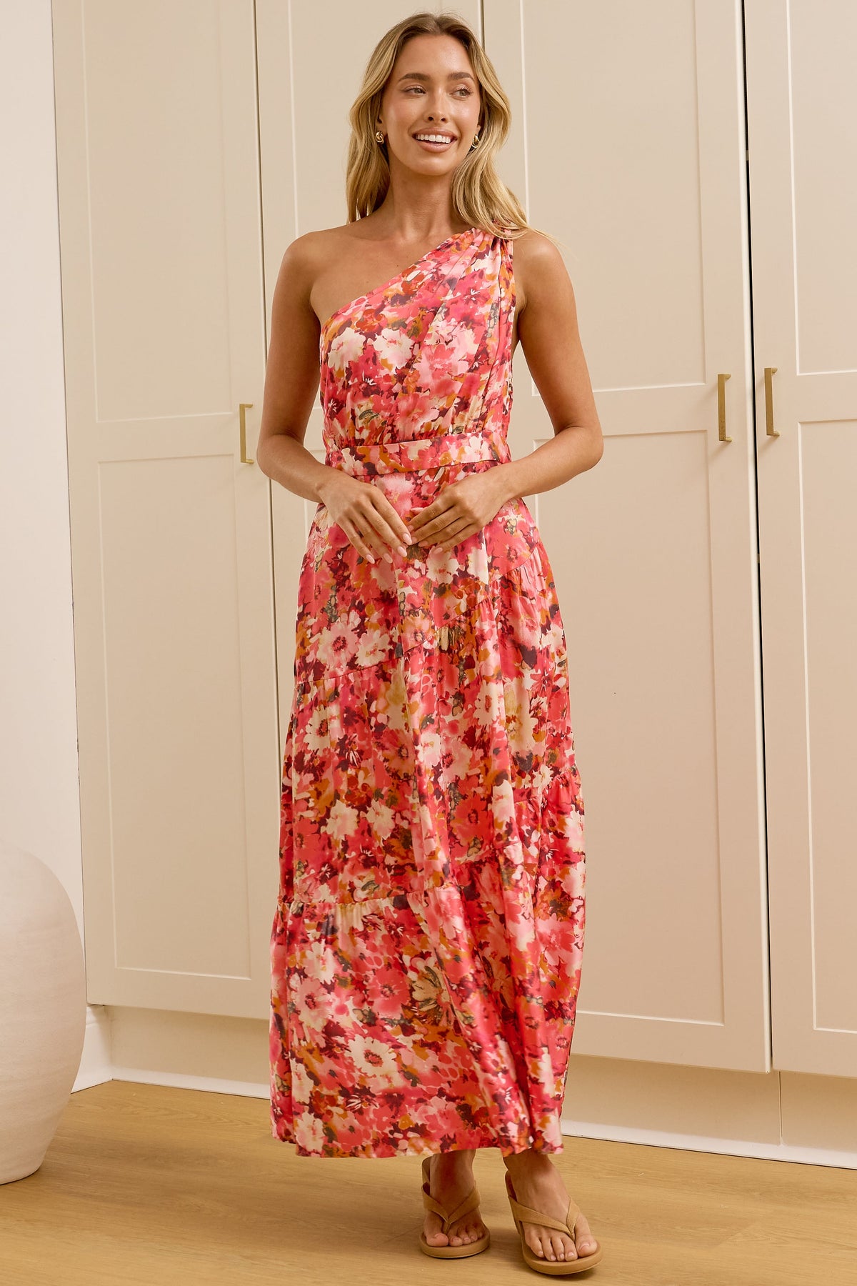 A woman stands in a bright room wearing a one-shoulder floral maxi dress with a red, pink, and orange pattern. She is positioned in front of a large gold-framed mirror that reflects her side profile, with tall cream-colored cabinets and two white ceramic vases in the background.