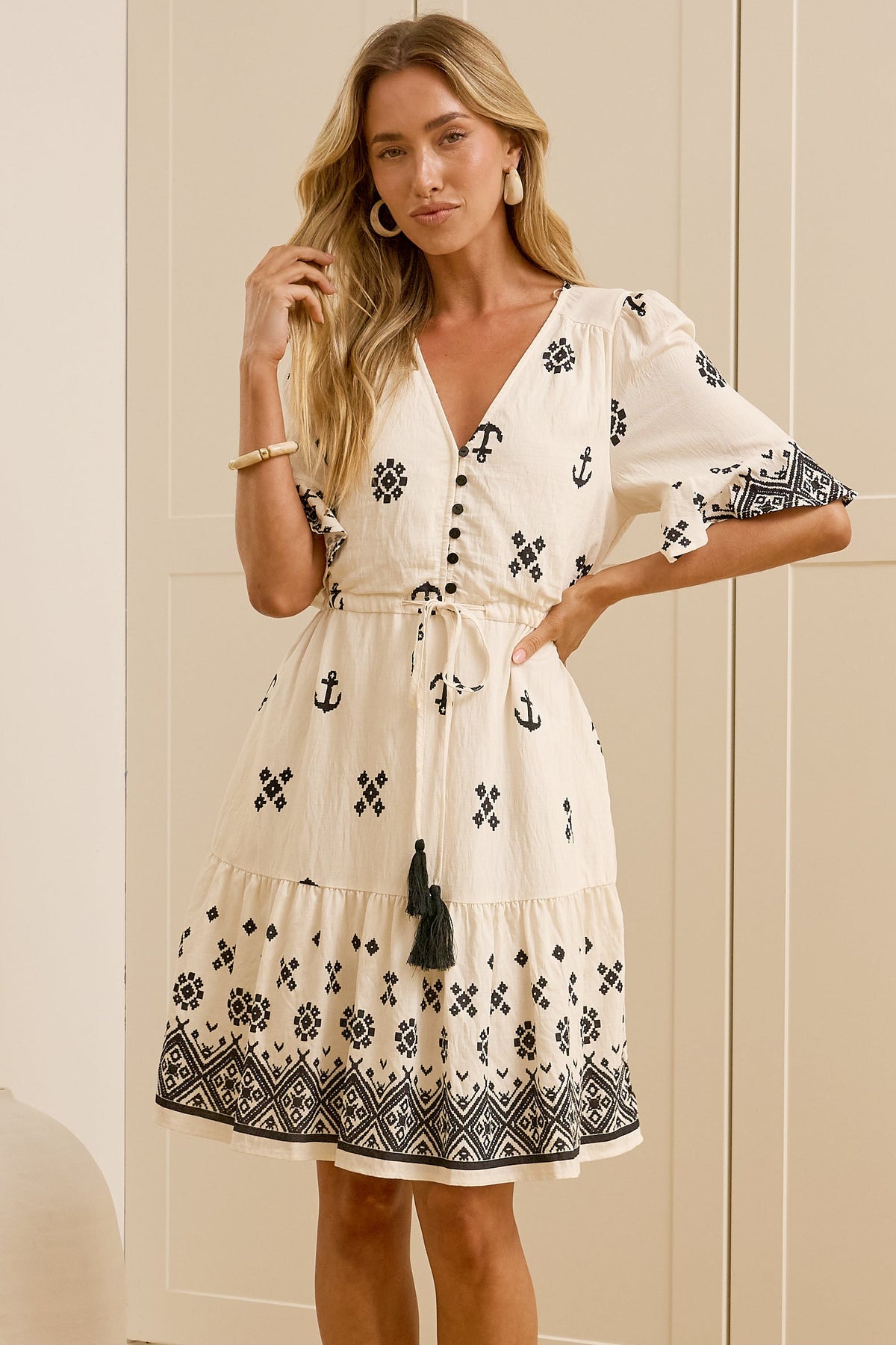 A woman stands in a bright room wearing a cream-colored dress with black geometric patterns and a matching tie belt. She is standing in front of white wardrobe doors and a large mirror that reflects her side profile.