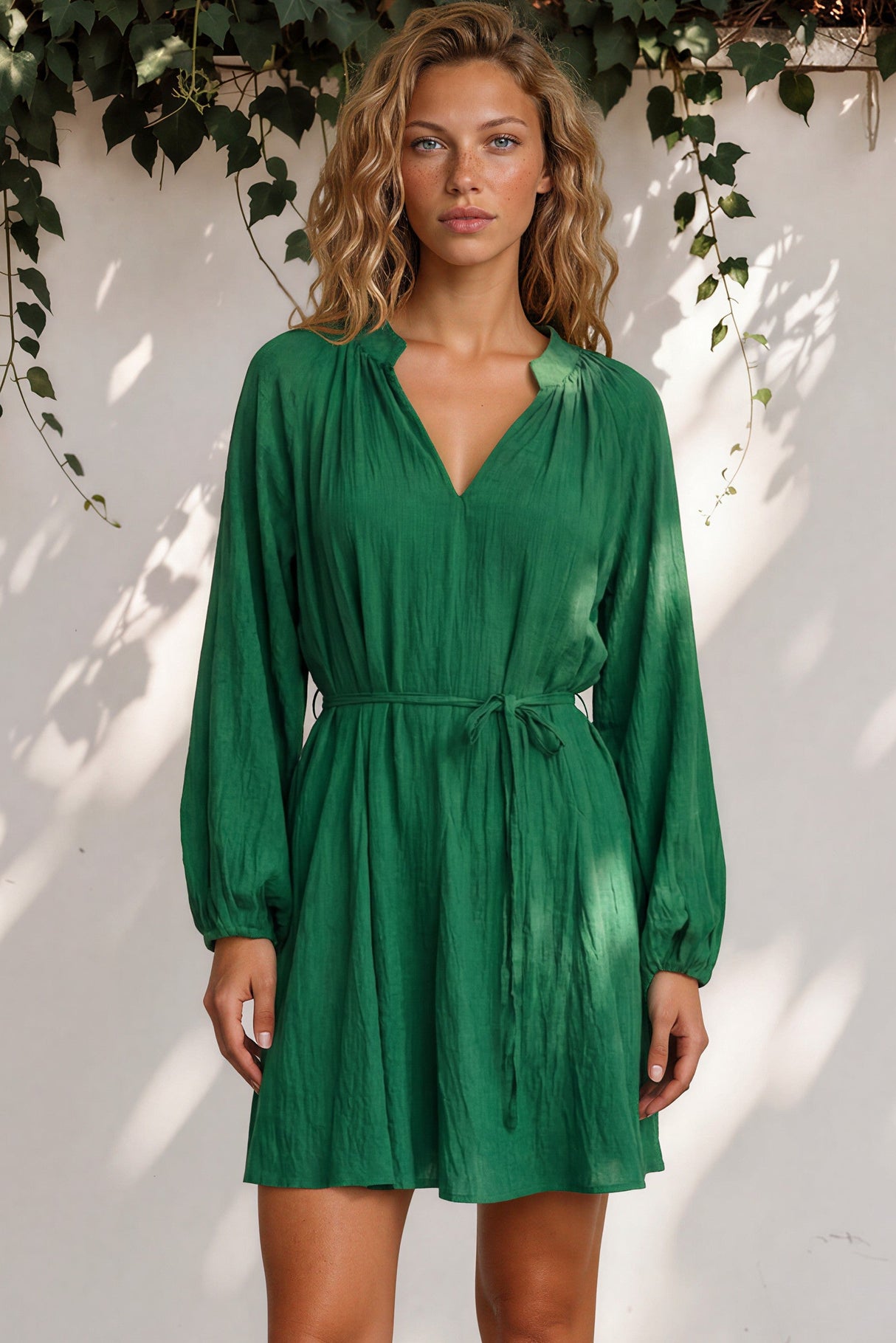 Woman wearing a green dress standing against a white wall with greenery.