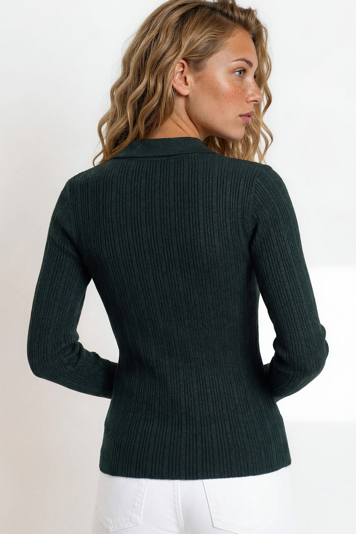 Woman wearing a dark green ribbed sweater and white pants against a light background