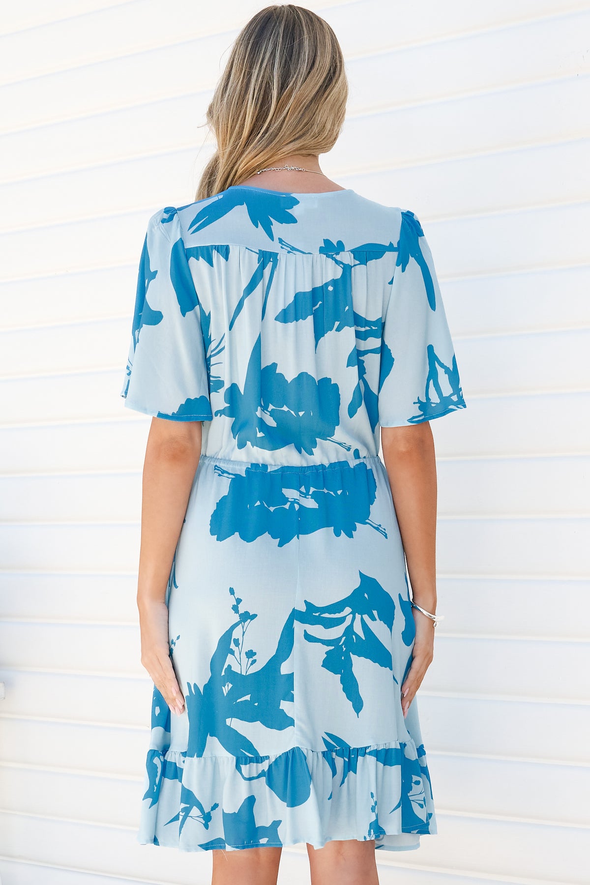 Back view of a woman wearing the Arabella Mini Dress in Colter Print Blue, showing the flutter sleeves, gathered tie waist and frill hem of the pale blue tropical leaf print dress, against a white wall