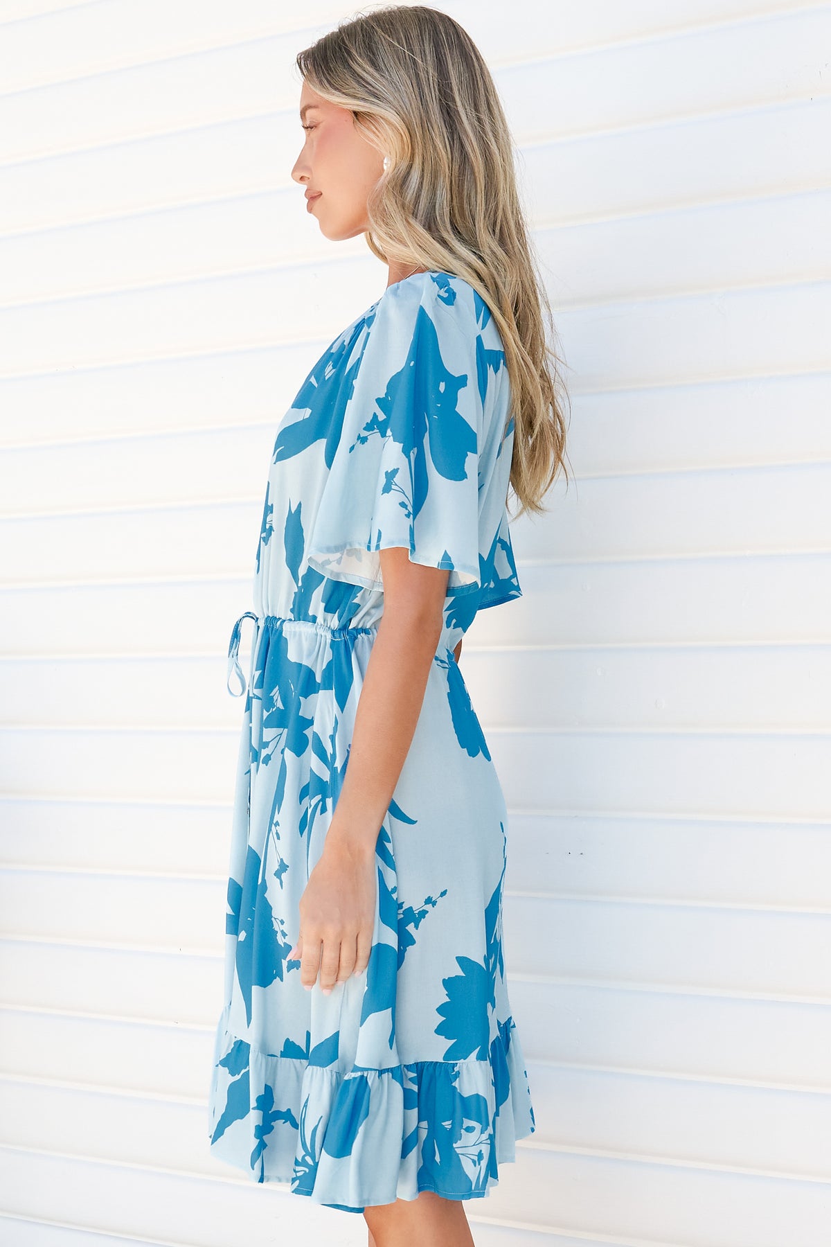 Side view of a woman wearing the Arabella Mini Dress in Colter Print Blue, showing the flutter sleeves, drawstring tie waist and frill hem of the pale blue tropical leaf print dress, against a white wall