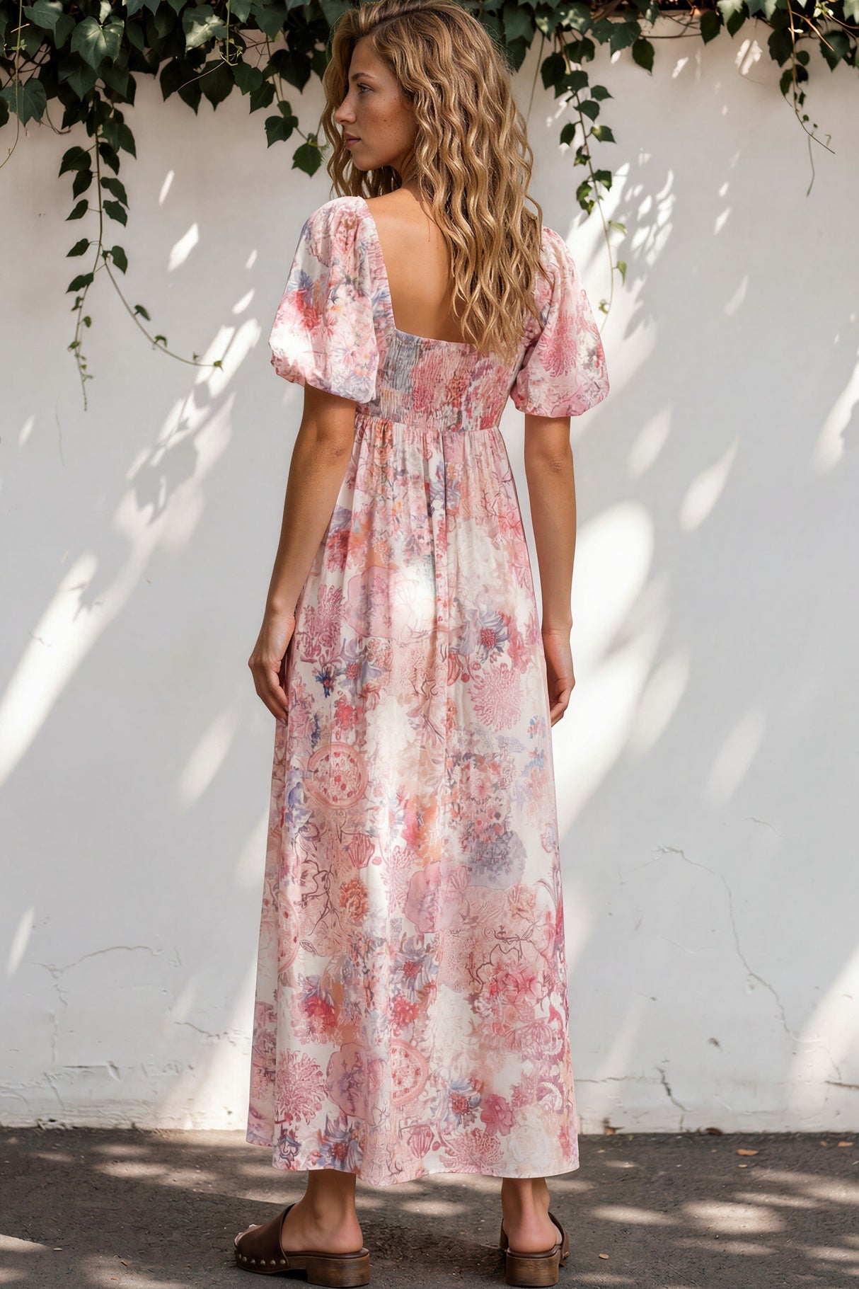 Woman wearing a floral dress standing against a white wall with greenery.