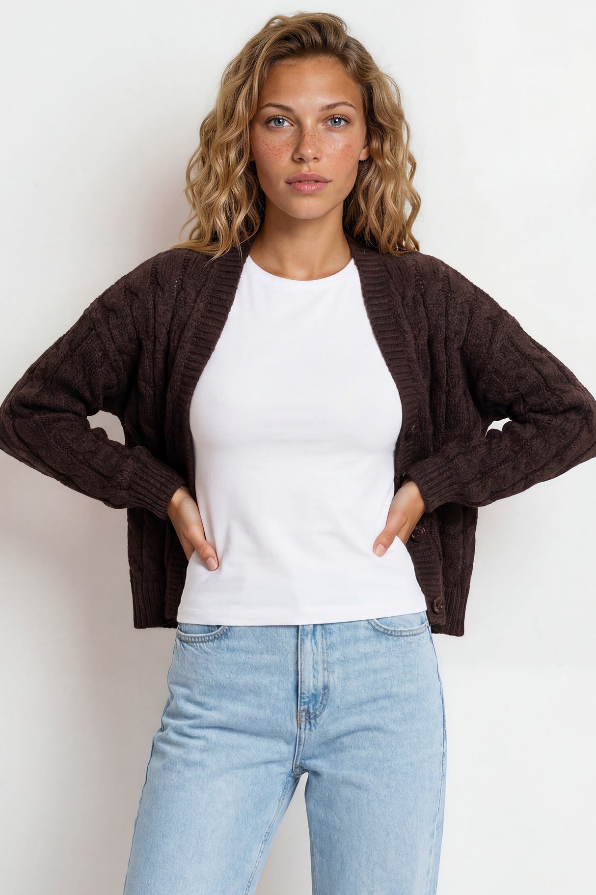 Woman wearing a brown cardigan, white shirt, and light blue jeans against a white background