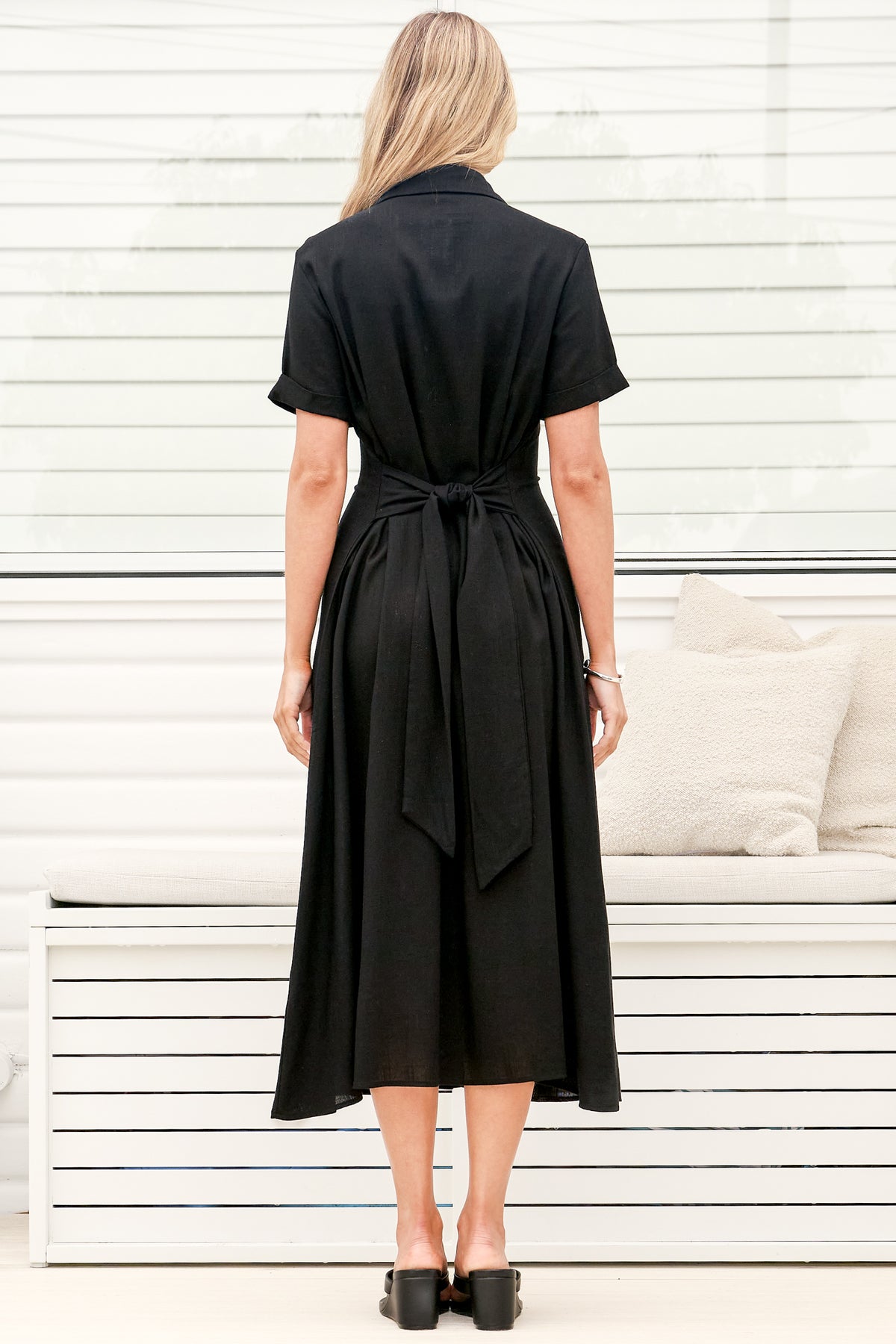 A woman wearing a black midi dress with a round neckline, short sleeves, and relaxed flowing silhouette, standing in front of a white panelled wall.
