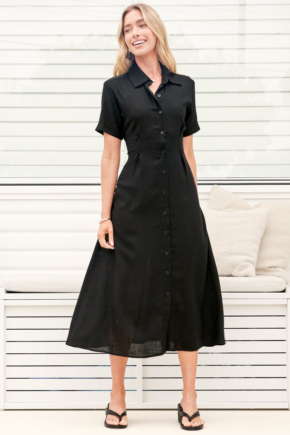 A woman wearing a black midi dress with a round neckline, short sleeves, and relaxed flowing silhouette, standing in front of a white panelled wall.