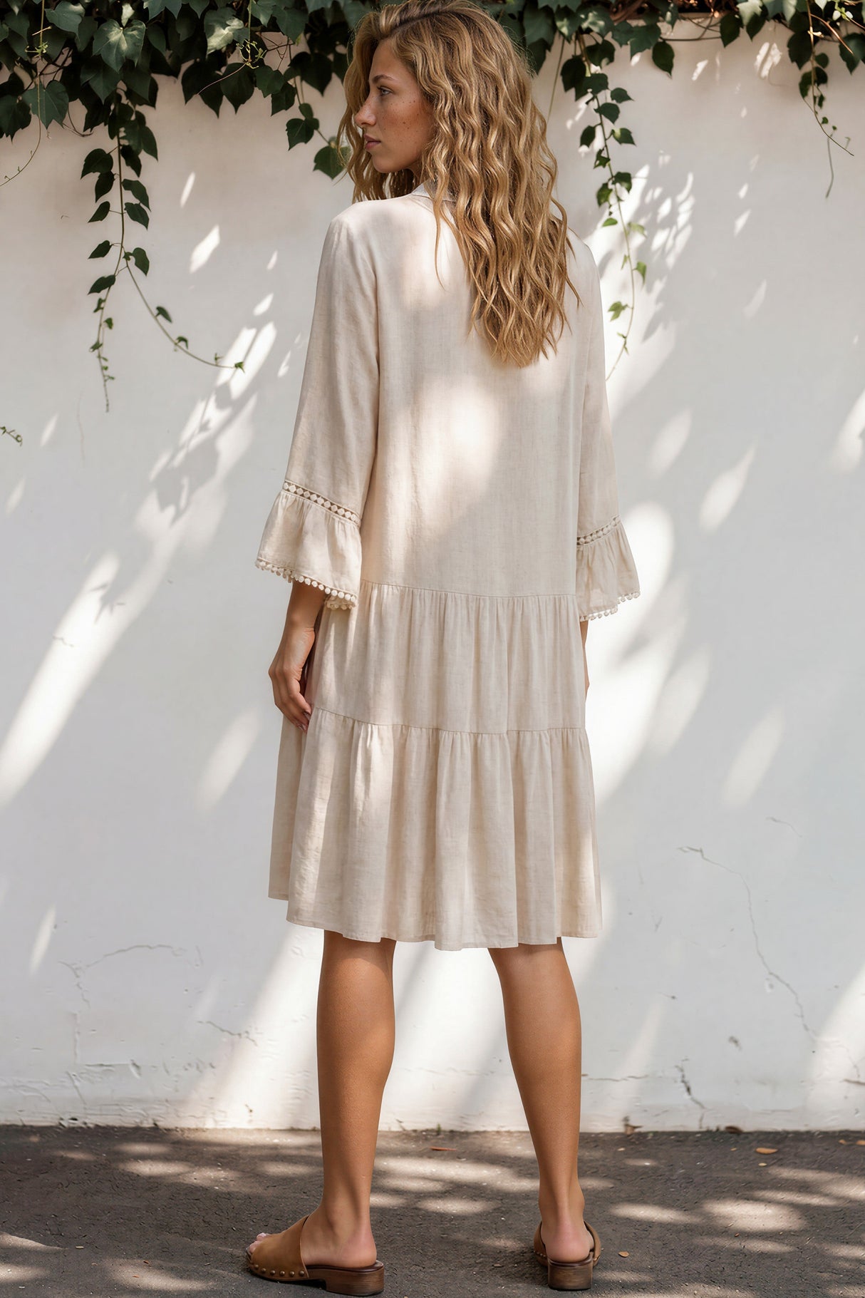 Woman wearing a beige dress standing against a white wall with greenery.