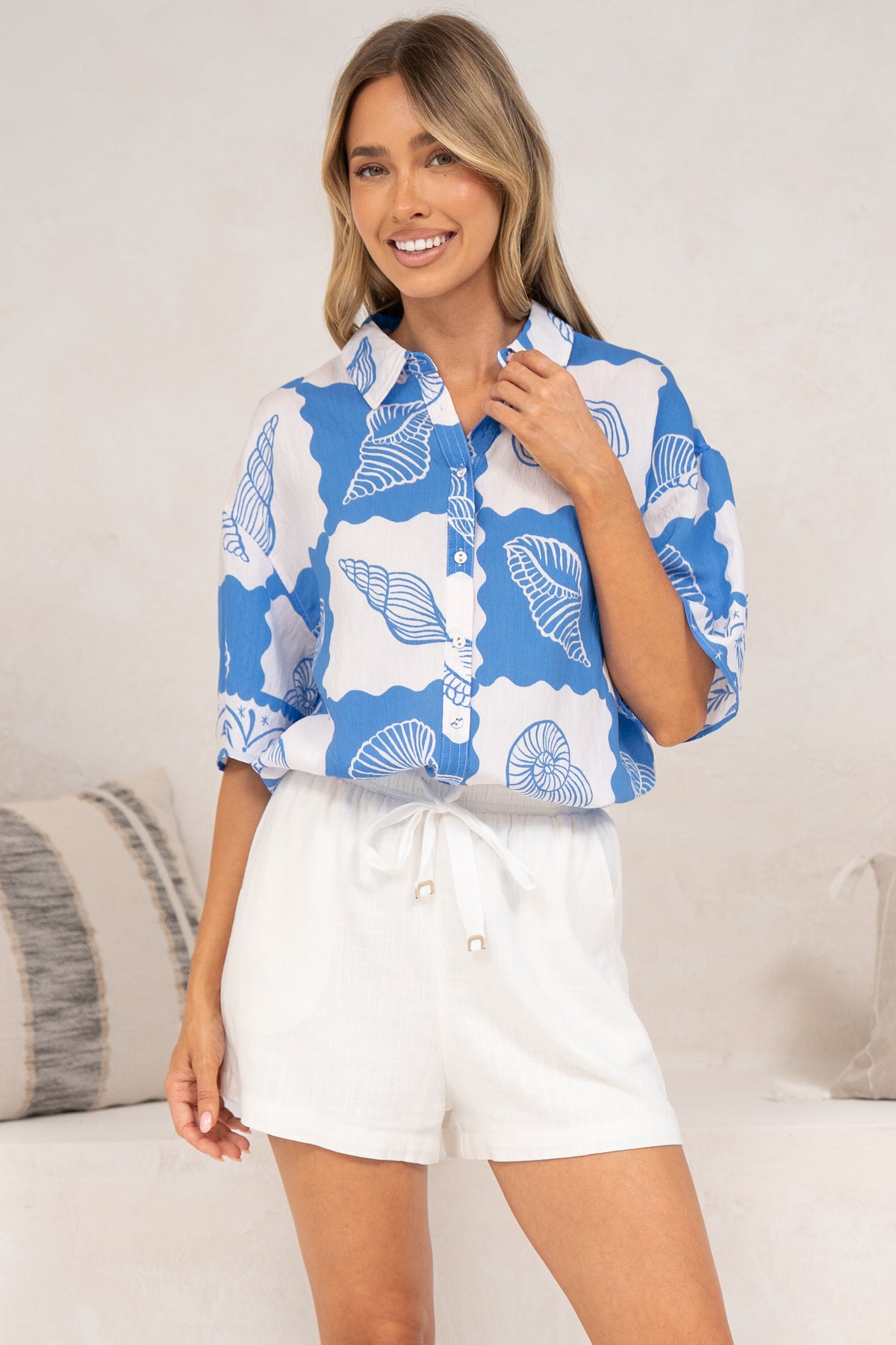 A smiling woman wearing the Blaze Shirt in Atrina Print tucked into white drawstring shorts, showing the collared short-sleeve button-down in a blue and white seashell print, in front of a cream sofa with neutral cushions