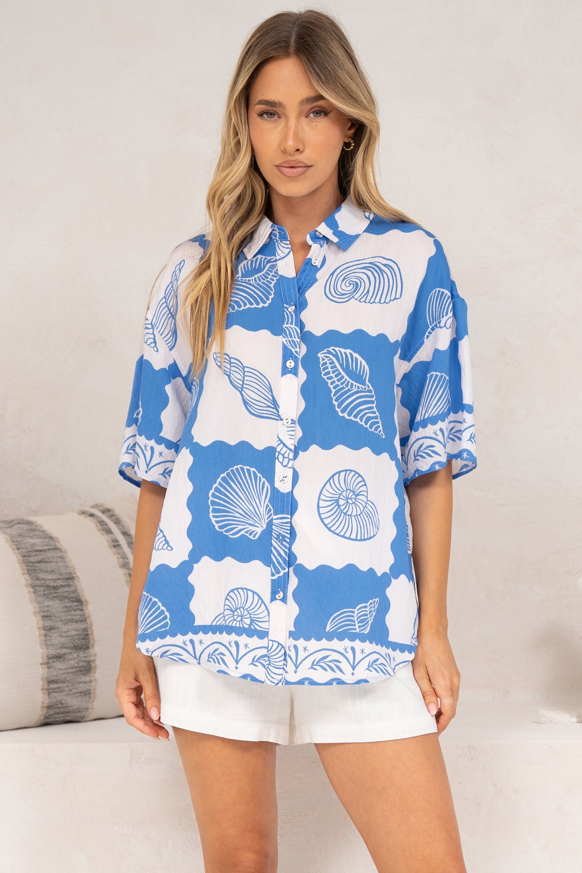 A woman standing front-on in the Blaze Shirt in Atrina Print, a relaxed collared short-sleeve button-down with a blue and white seashell print worn loose over white shorts, in front of a cream sofa with neutral cushions