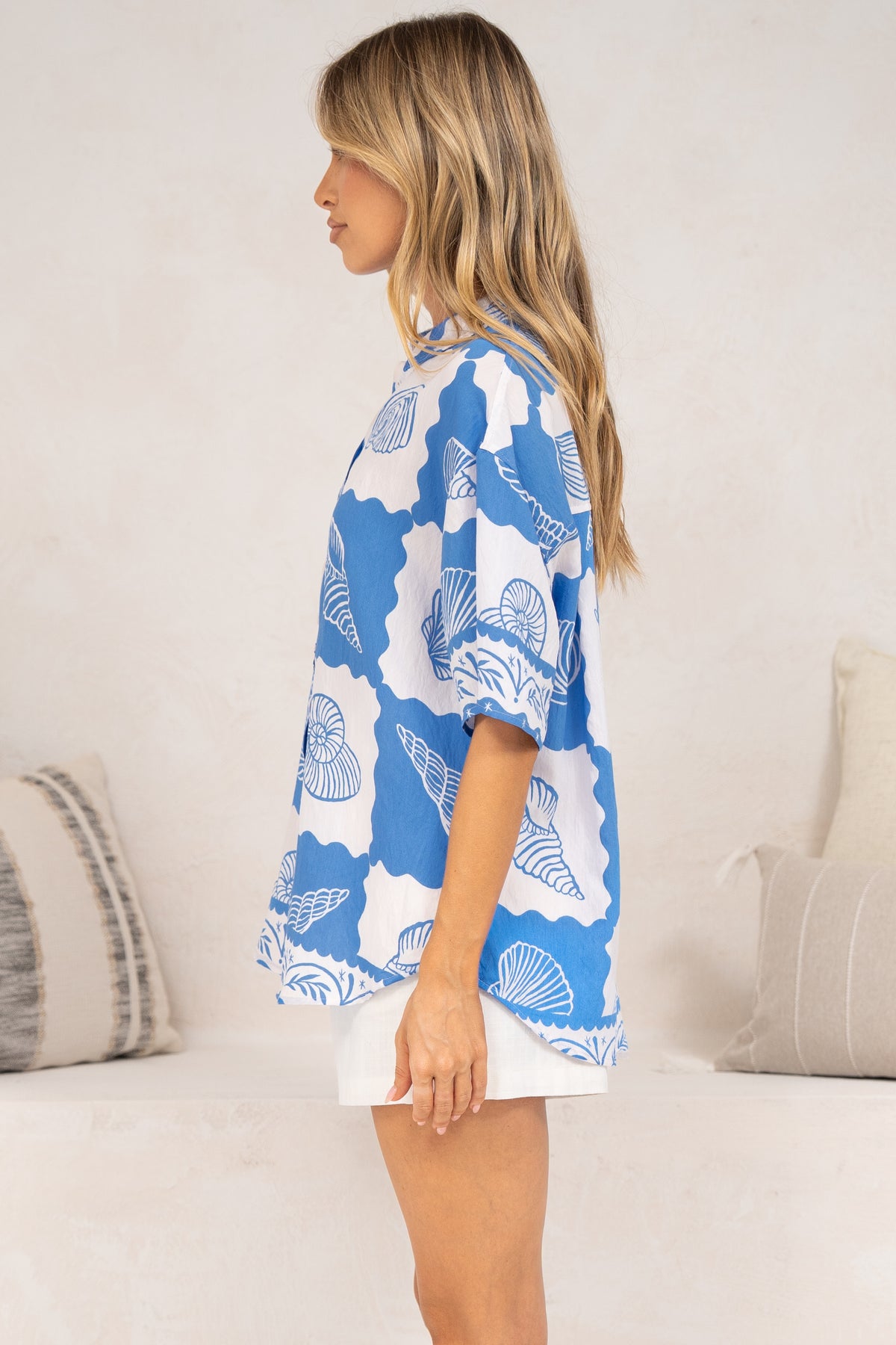 Side view of a woman wearing the Blaze Shirt in Atrina Print, showing the relaxed oversized fit and short sleeves of the blue and white seashell print button-down, styled with white shorts in front of a cream sofa
