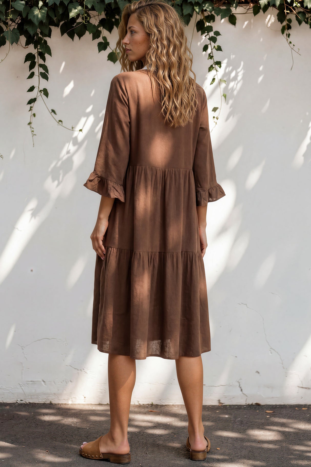 Woman wearing a brown dress standing against a white wall with greenery.
