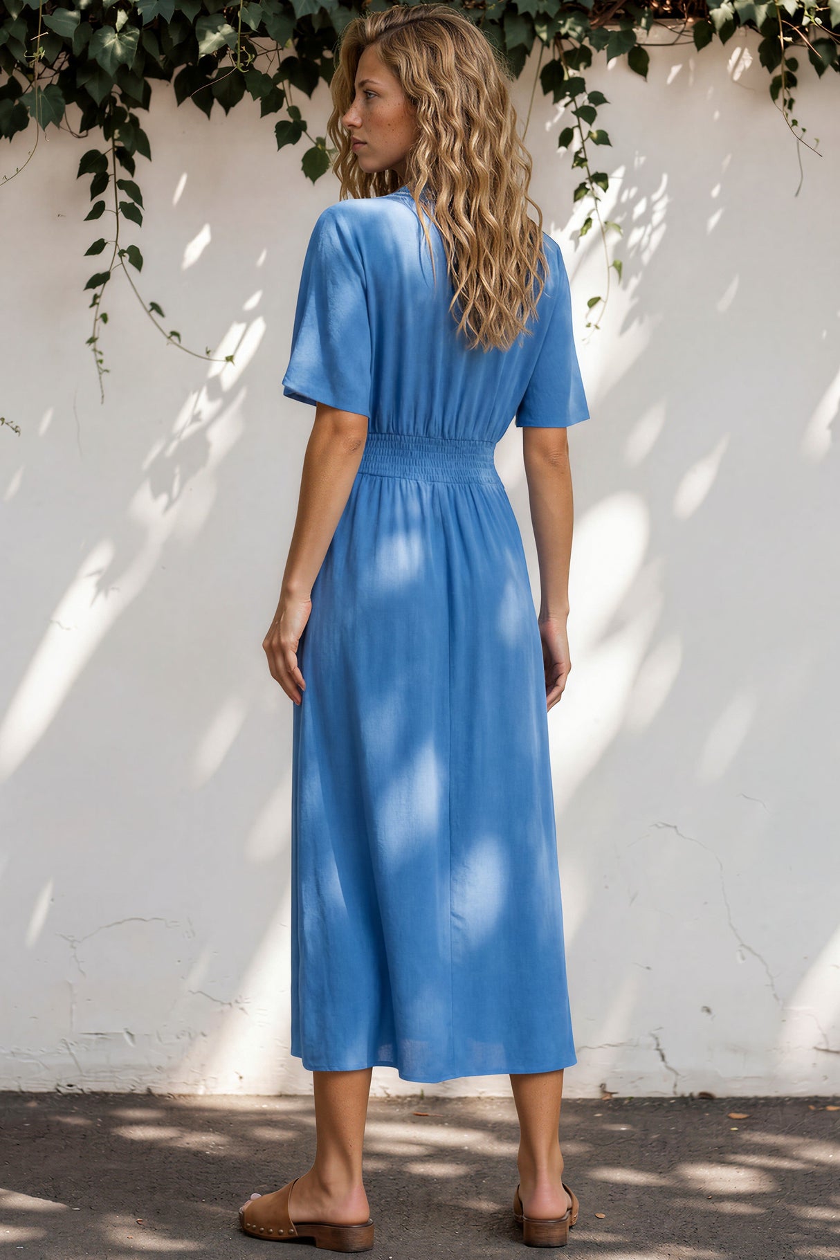 Woman in a blue dress standing against a white wall with greenery.