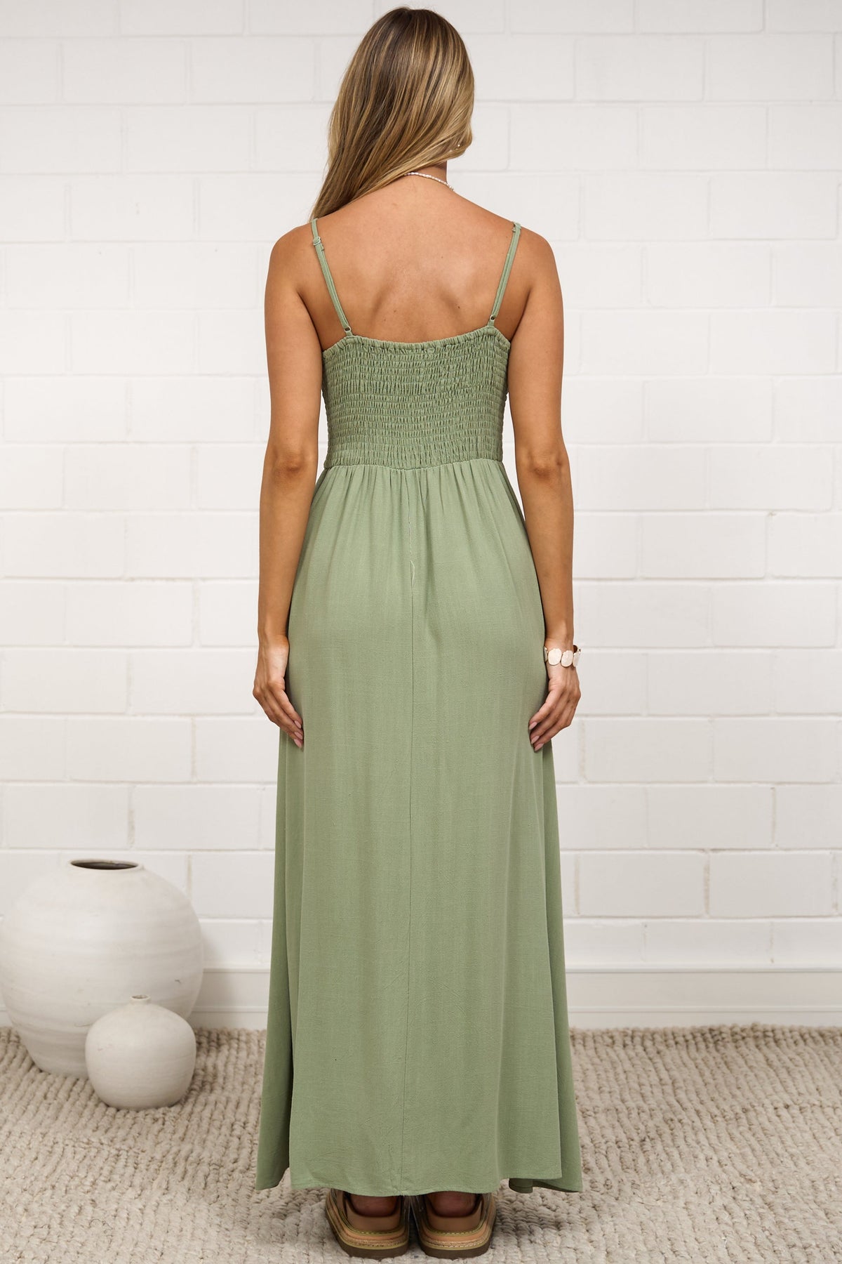 A woman stands with her back to the camera wearing a strapless sage green maxi dress in a bright, modern room with a white brick wall and neutral decor.