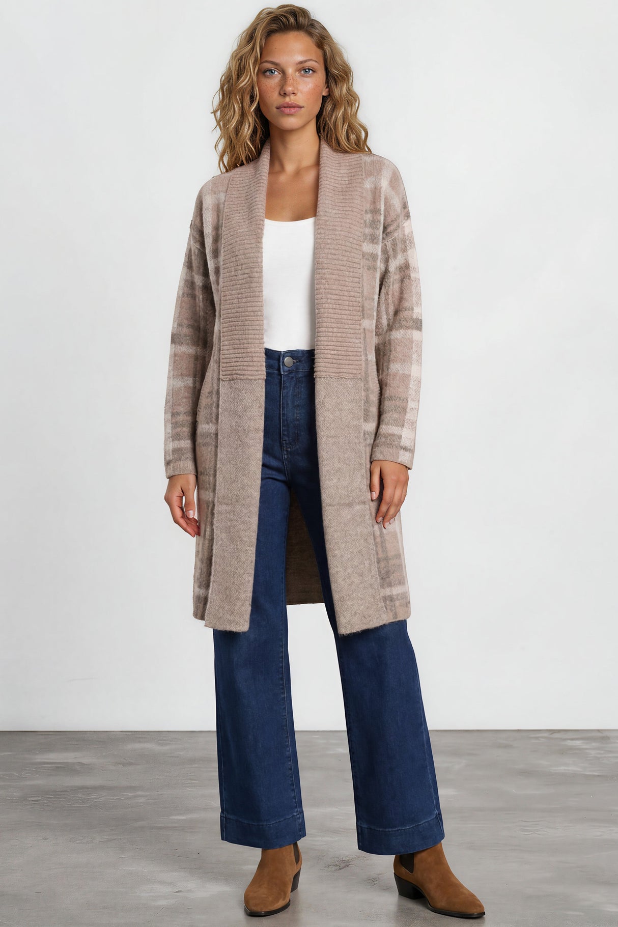 Woman wearing a long beige cardigan, white top, blue jeans, and brown shoes on a plain background