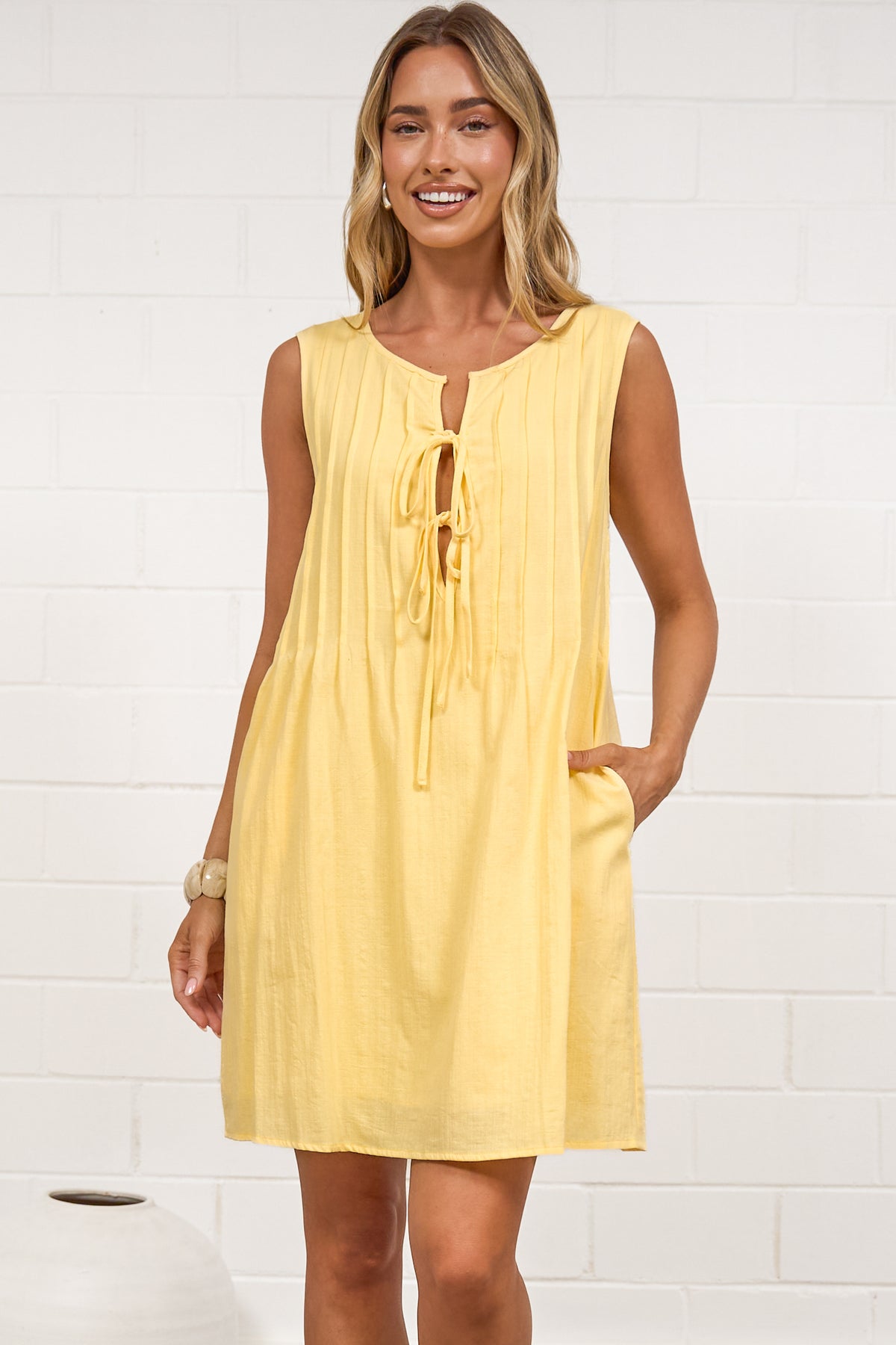 A woman in a bright yellow sleeveless mini dress featuring a keyhole neckline, pintuck details, and side pockets, posed in front of a white wall.