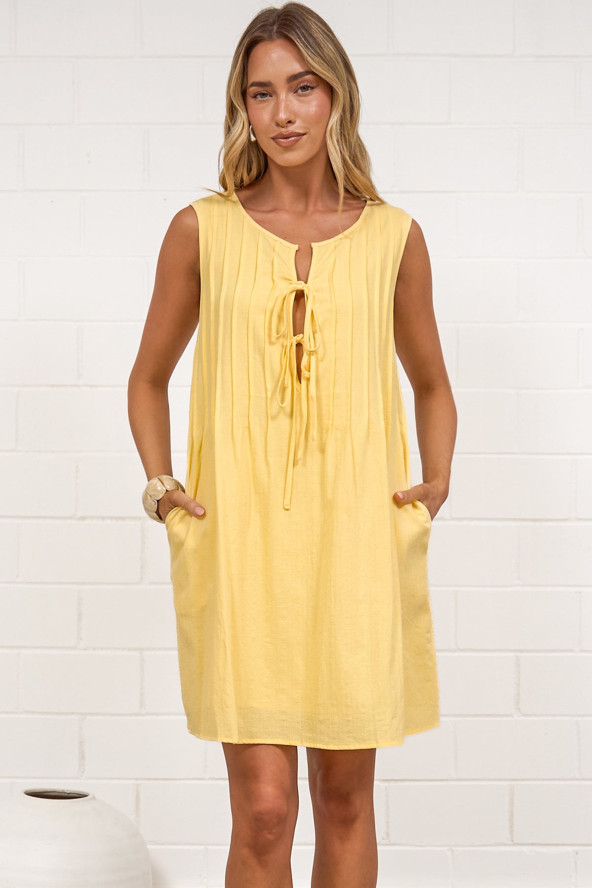 A woman in a bright yellow sleeveless mini dress featuring a keyhole neckline, pintuck details, and side pockets, posed in front of a white wall.