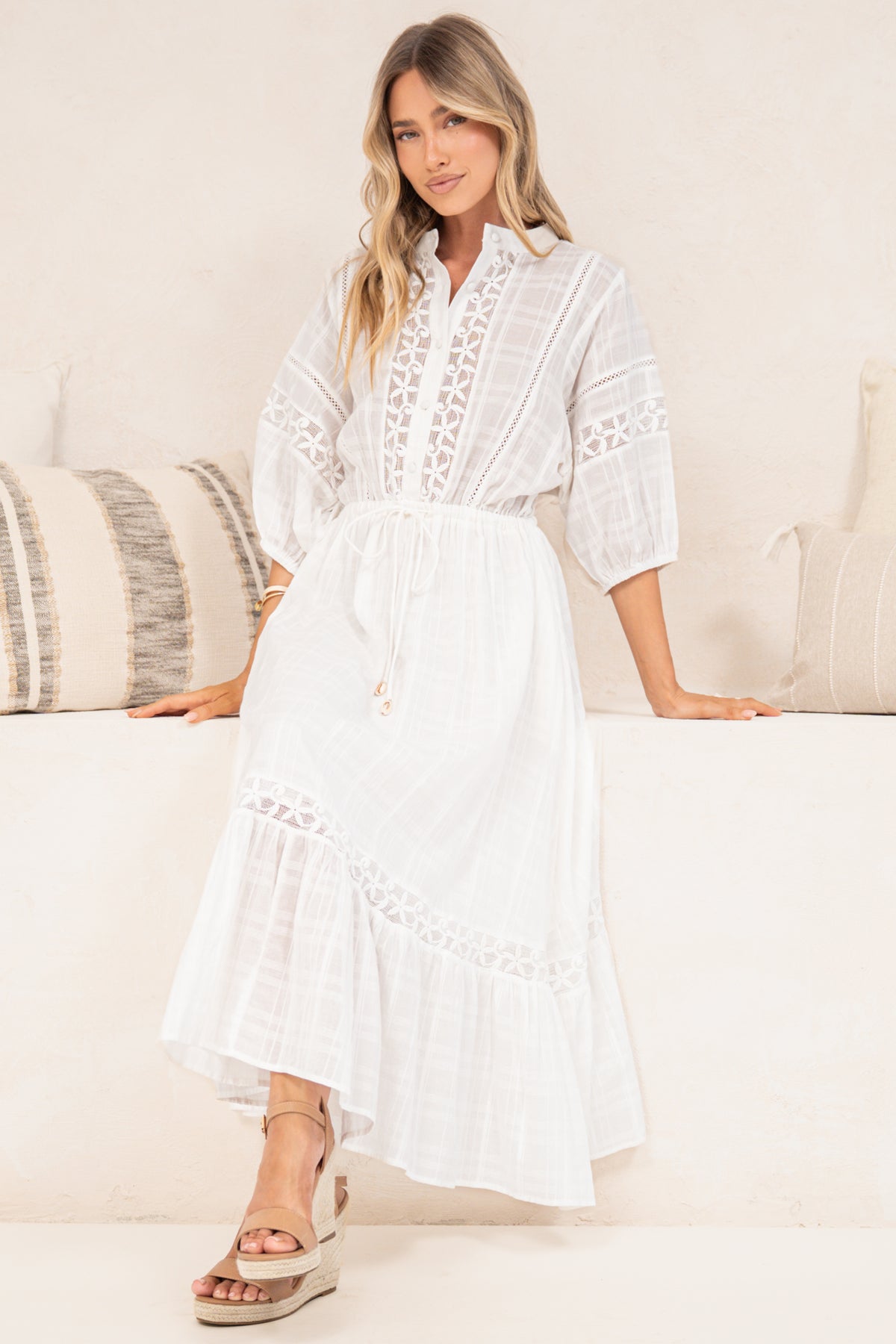 A woman wearing a white midi dress with intricate floral lace embroidery and a drawstring waist, leaning against a white wall in a brightly lit indoor setting.