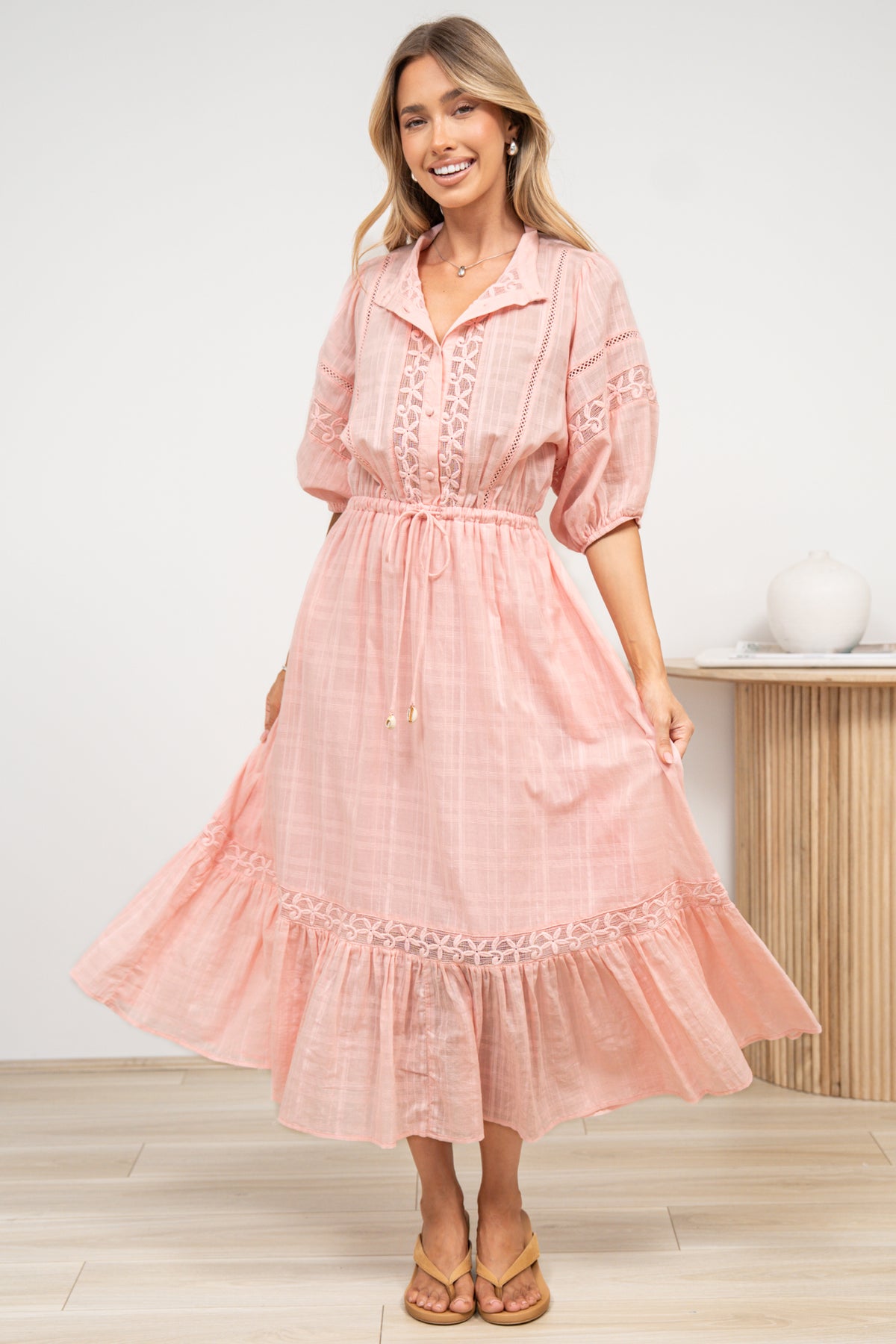 A woman in a romantic pink maxi dress featuring a sheer plaid texture, lace detailing, and flowing fluted hem, posed against an off-white painted wall.