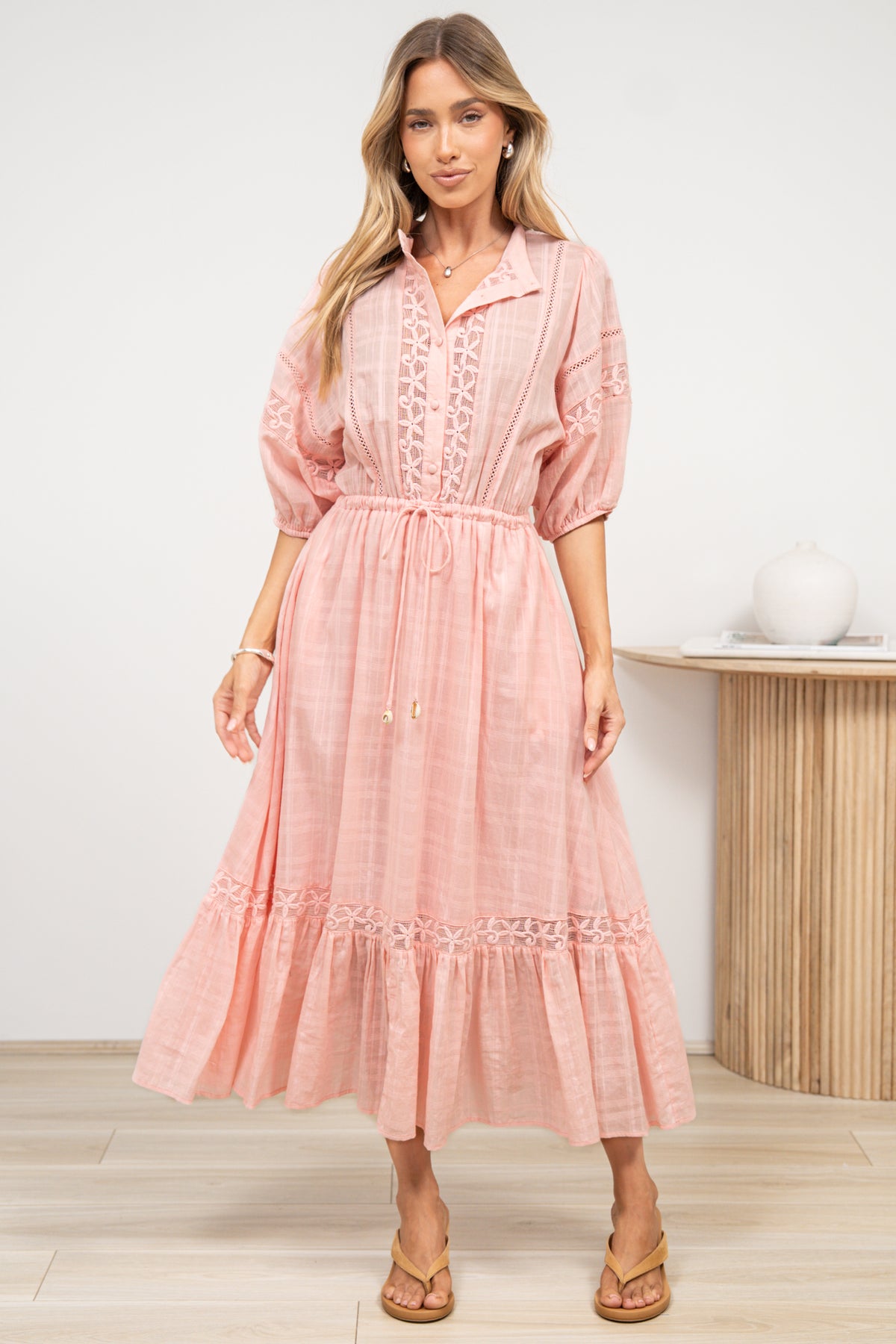 A woman in a romantic pink maxi dress featuring a sheer plaid texture, lace detailing, and flowing fluted hem, posed against an off-white painted wall.