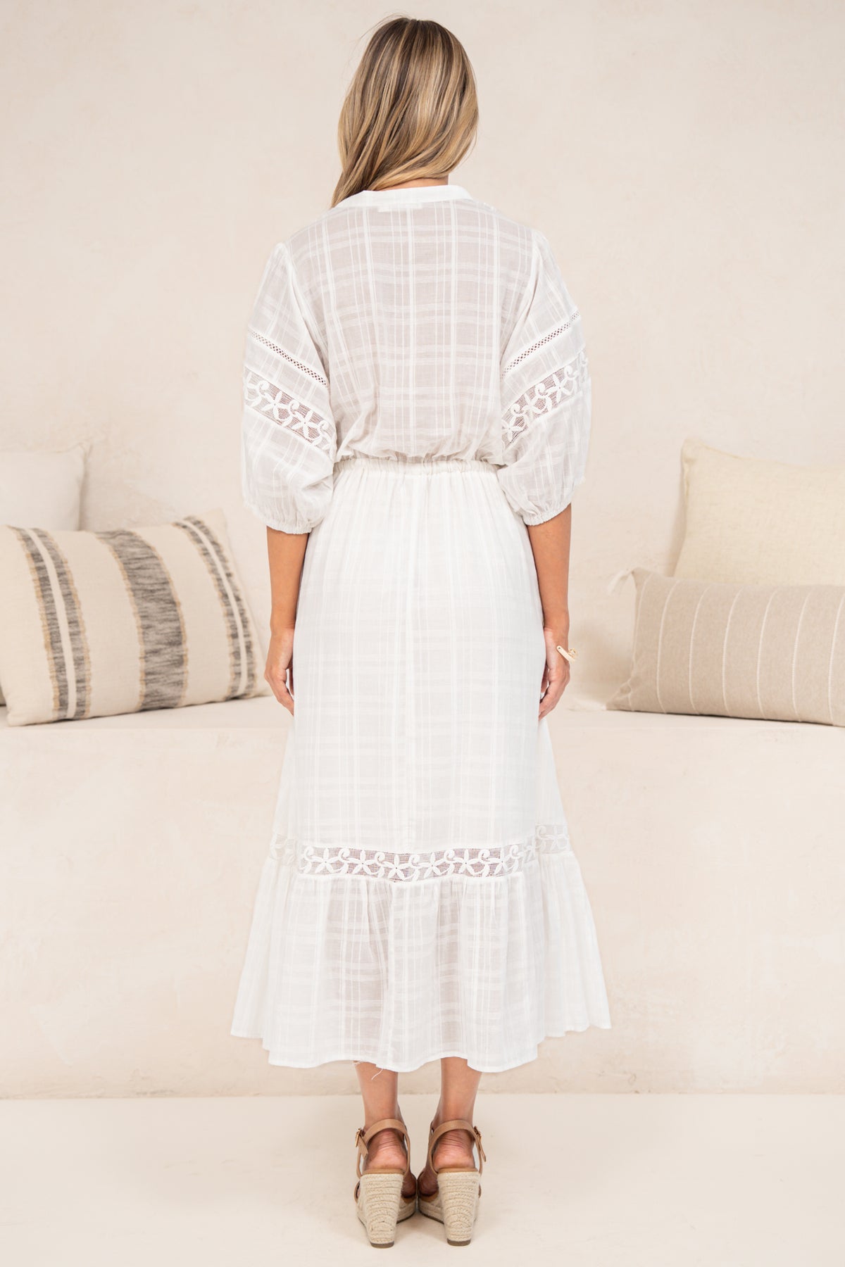 A woman in a flowing white maxi dress featuring sheer plaid-textured fabric, delicate lace detailing, and puff sleeves, posed beside a cream couch.
