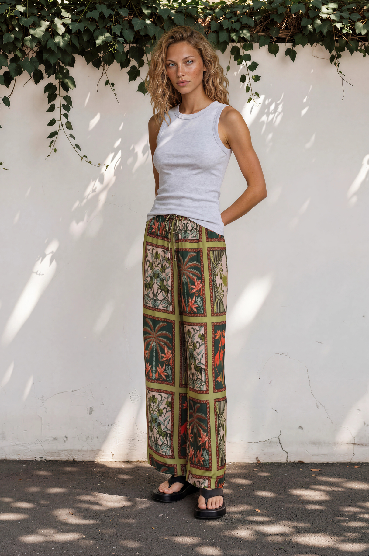 Woman wearing a white sleeveless top and patterned wide-leg pants standing against a white wall with greenery.