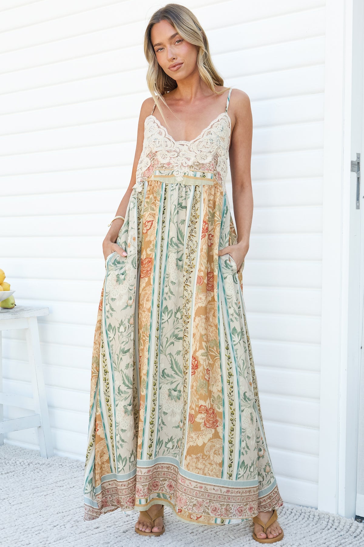 A woman with her hands in her pockets wearing the Georgette Maxi Dress in Lyla Print, a relaxed floor-length sun dress with a lace V-neck bodice, thin straps and a cream and sage floral stripe print, against a white wall