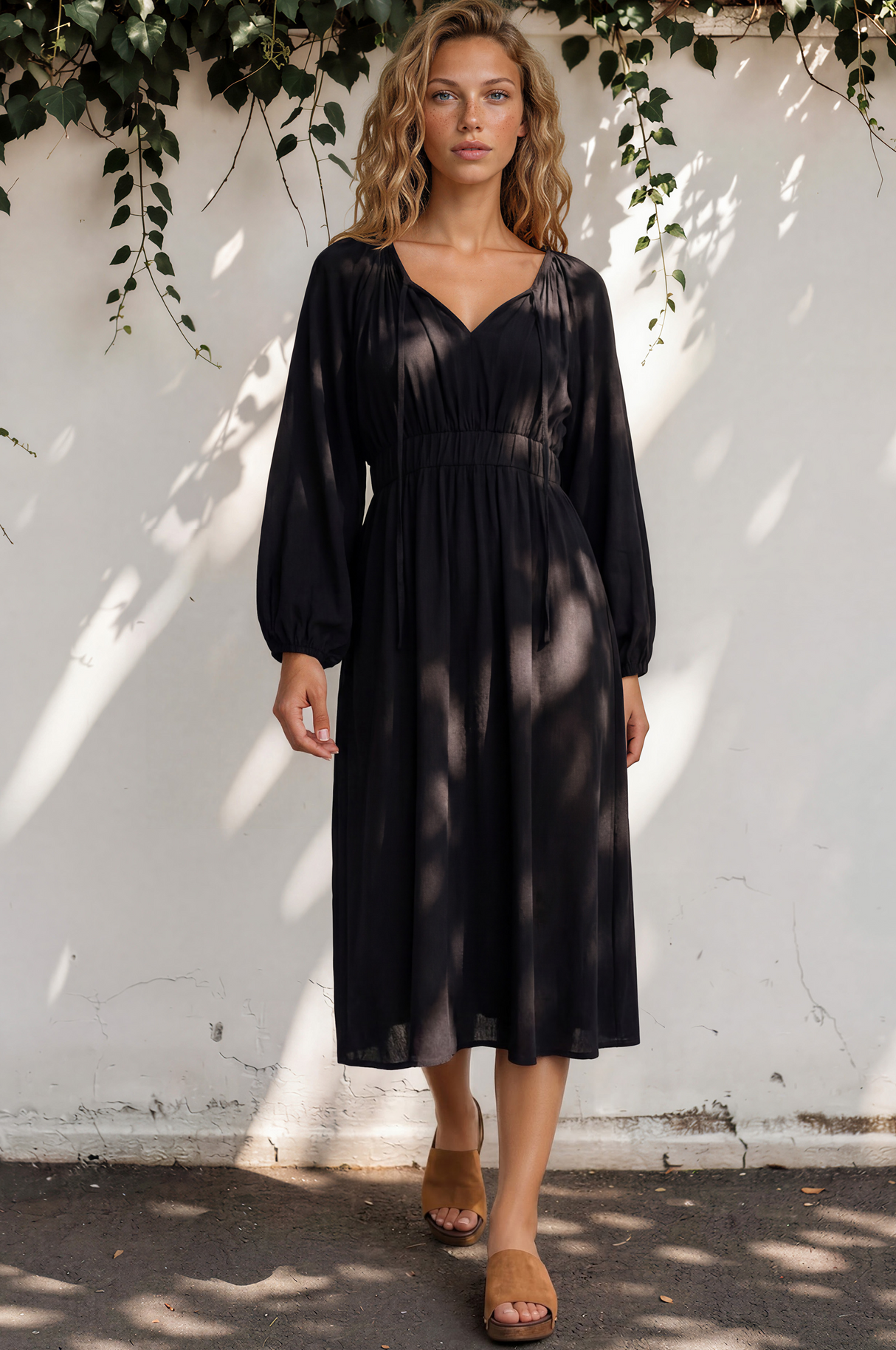 Woman wearing a black dress standing against a white wall with greenery.