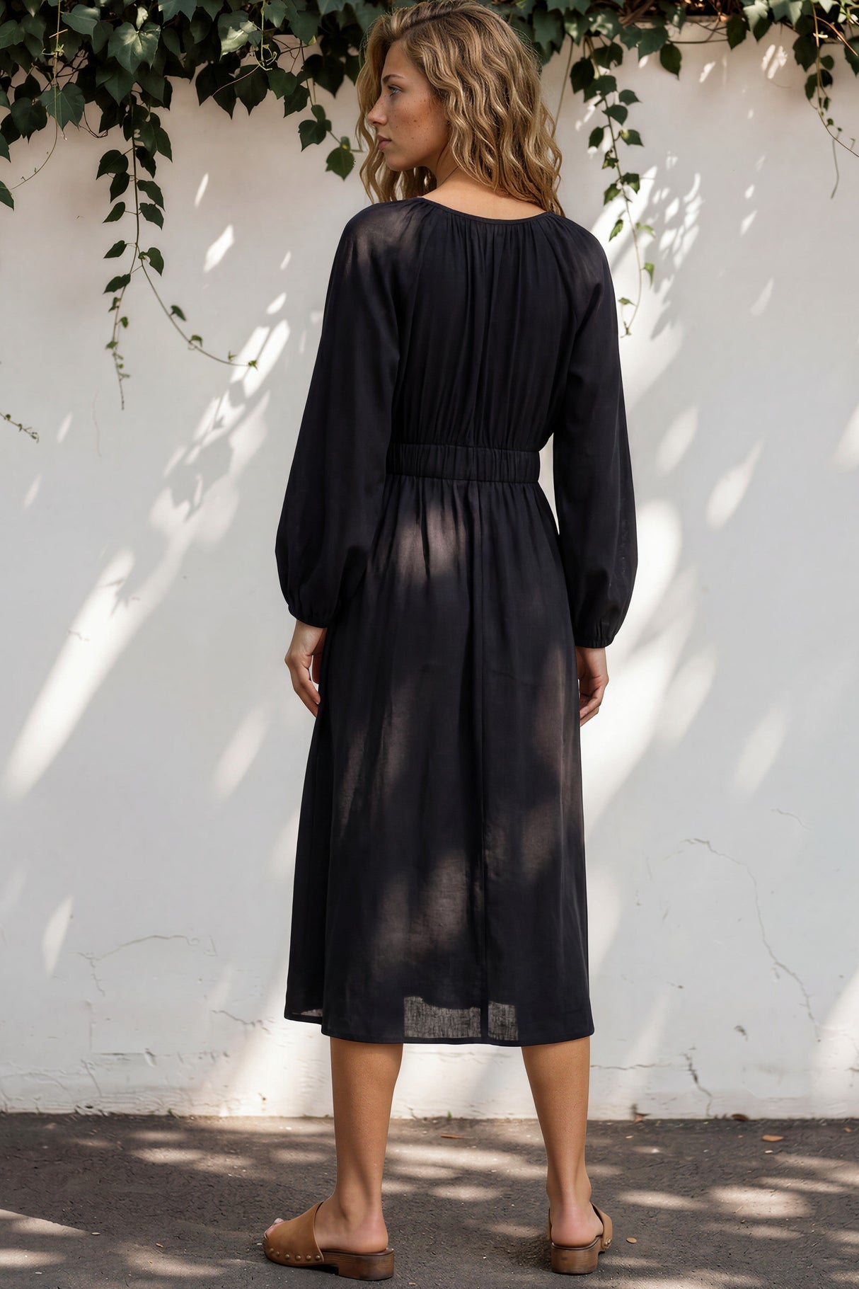 Woman wearing a black dress standing against a white wall with greenery.