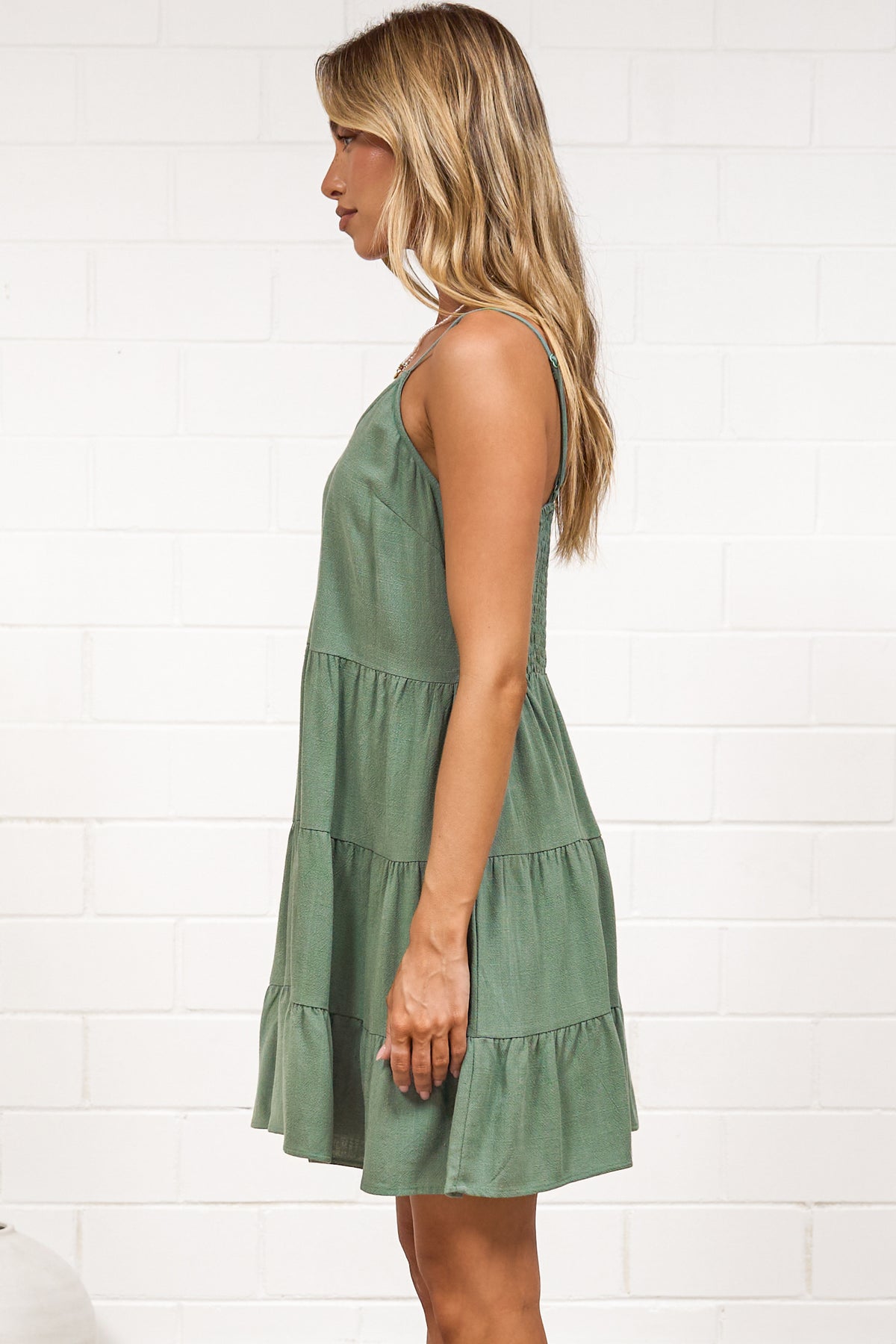 A woman in a dark green mini dress with a V-neckline, adjustable spaghetti straps, and a tiered ruffled hem, posed against a white painted brick wall.