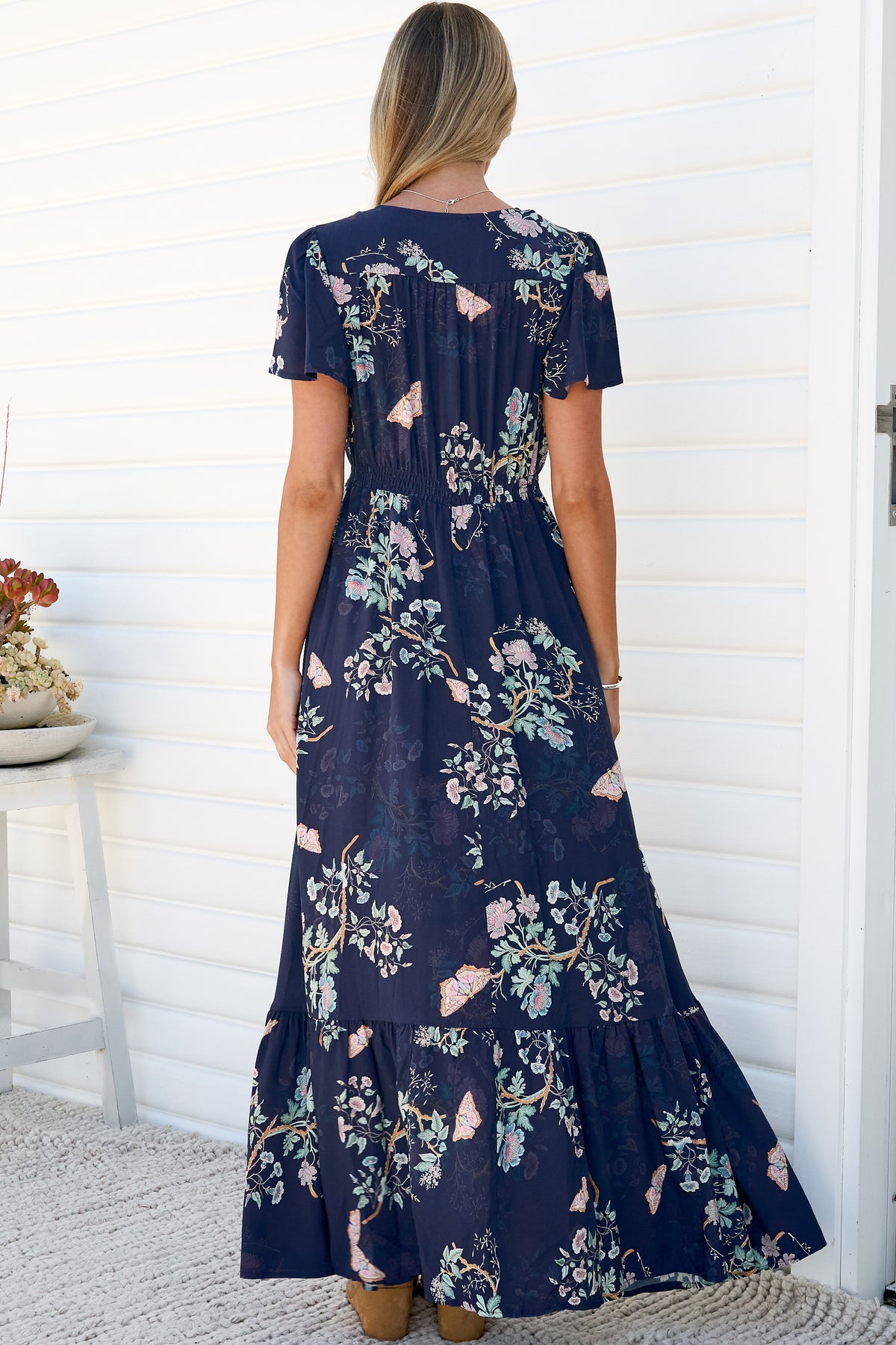 Back view of a woman wearing the Kali Maxi Dress in Colette Print, showing the flutter sleeves, smocked waist and tiered floor-length skirt of the navy floral maxi dress, against a white wall