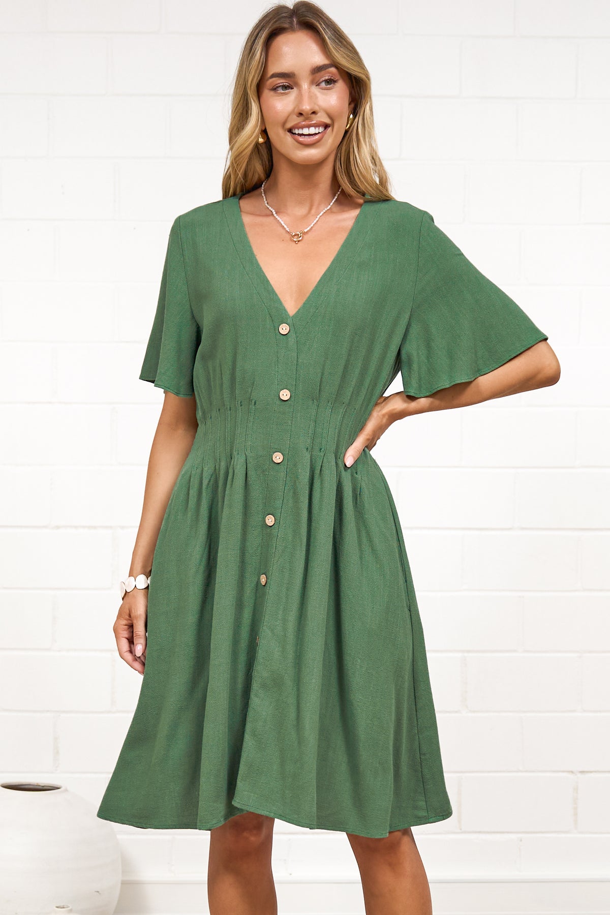 A woman in a green mini dress with a V-neckline, flutter sleeves, and decorative button placket, posed against a white painted brick wall.