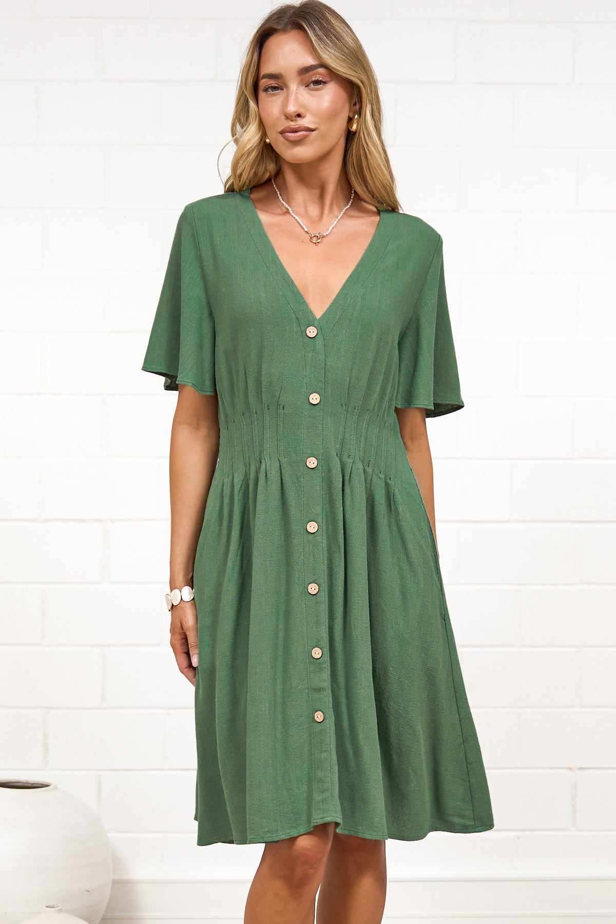 A woman in a green mini dress with a V-neckline, flutter sleeves, and decorative button placket, posed against a white painted brick wall.