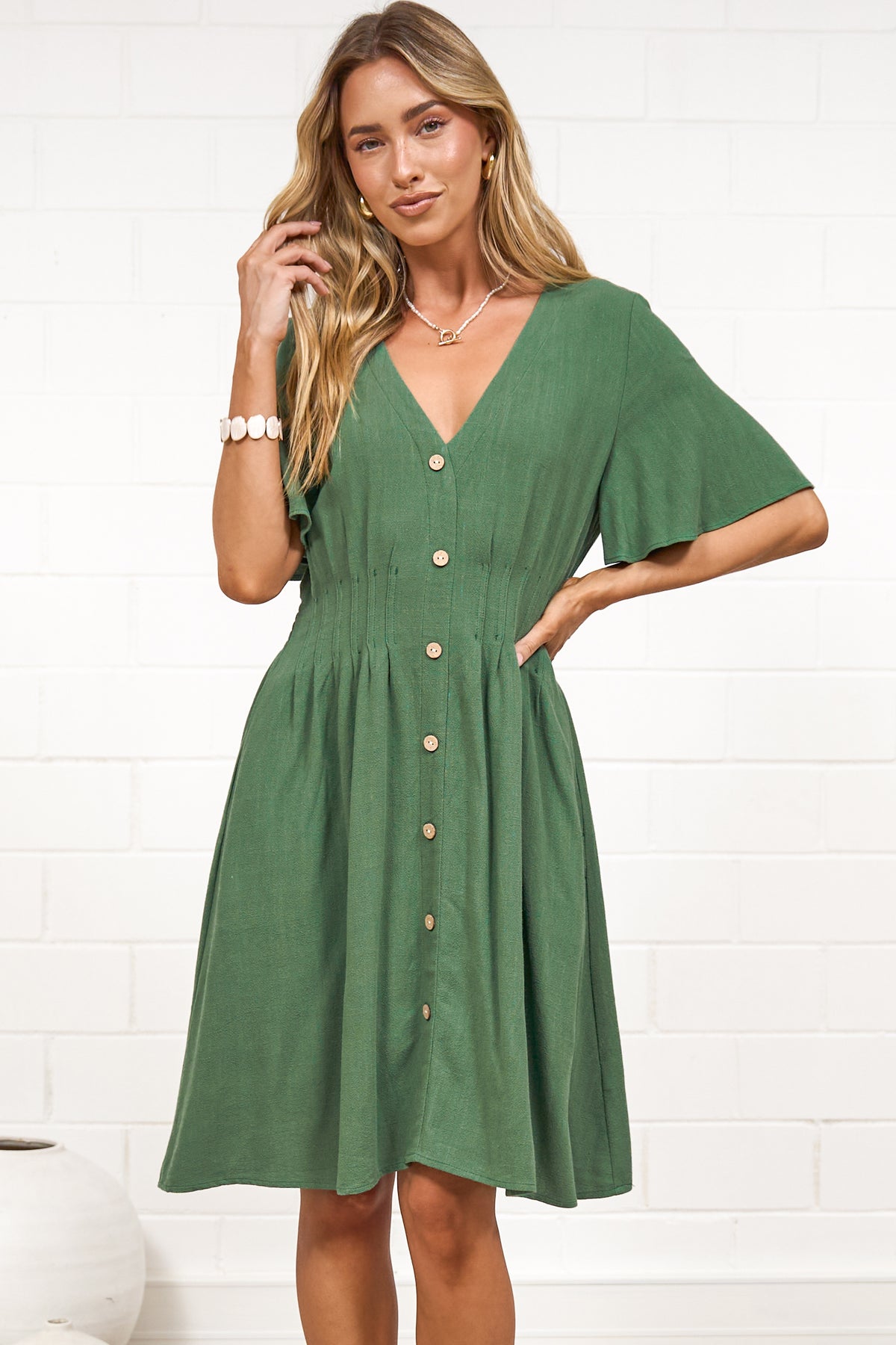 A woman in a green mini dress with a V-neckline, flutter sleeves, and decorative button placket, posed against a white painted brick wall.