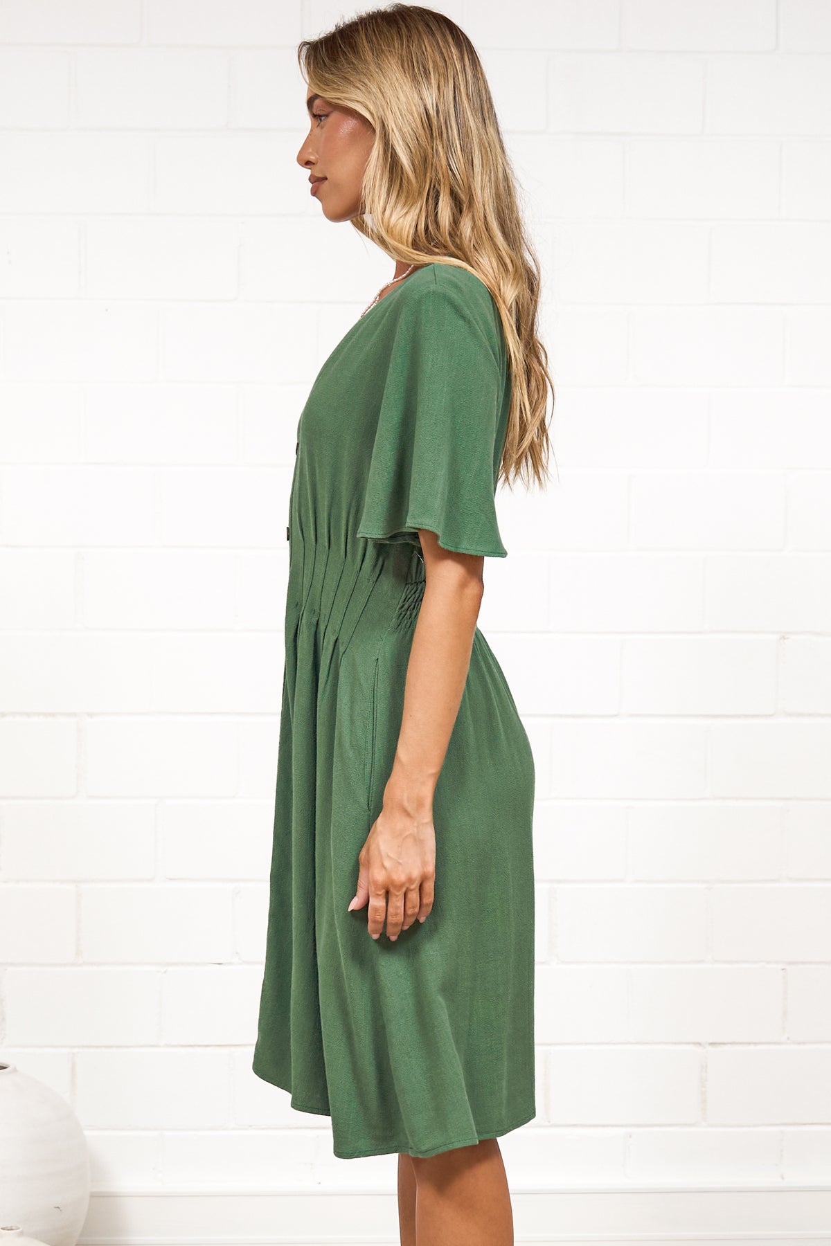 A woman in a green mini dress with a V-neckline, flutter sleeves, and decorative button placket, posed against a white painted brick wall.