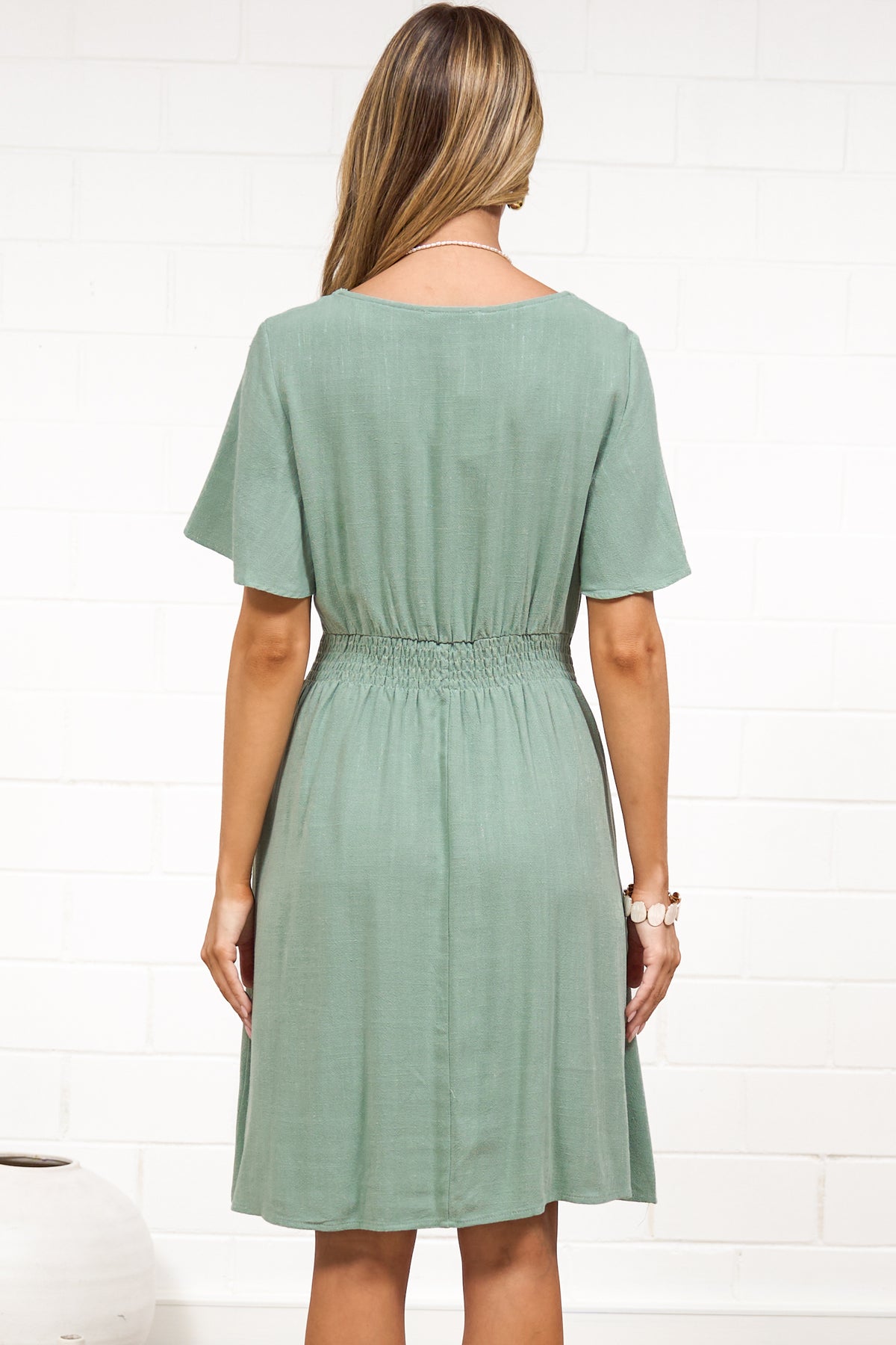 A woman in a sage mini dress with flutter sleeves, a V-neckline, and decorative button placket, posed against a white painted brick wall.