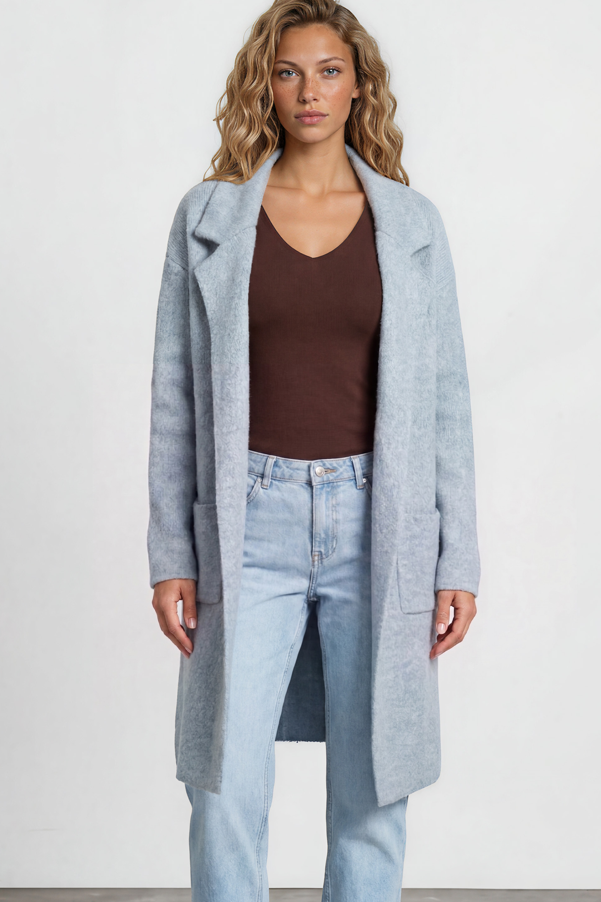 Woman wearing a long light blue coat over a brown top and jeans on a white background