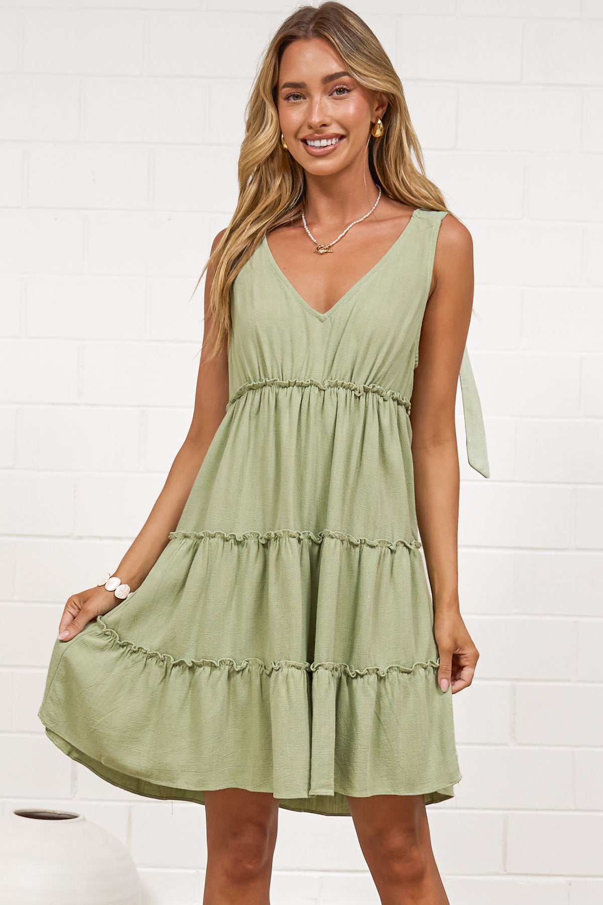 A woman wearing a sage mini dress with tie shoulder straps, a soft V-neckline, tiered skirt, and frill detailing, standing against a white painted brick wall.