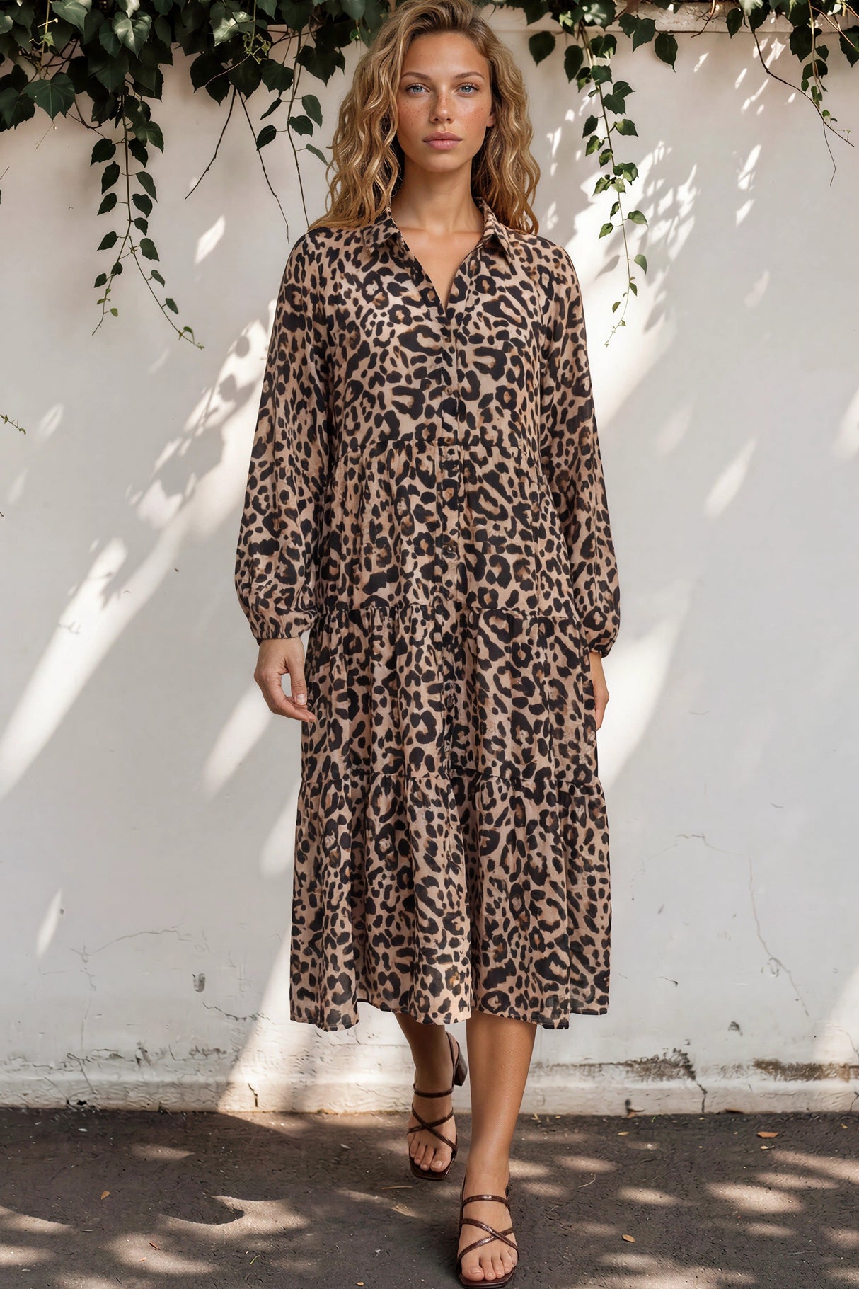 Woman wearing a leopard print dress standing against a white wall with greenery.