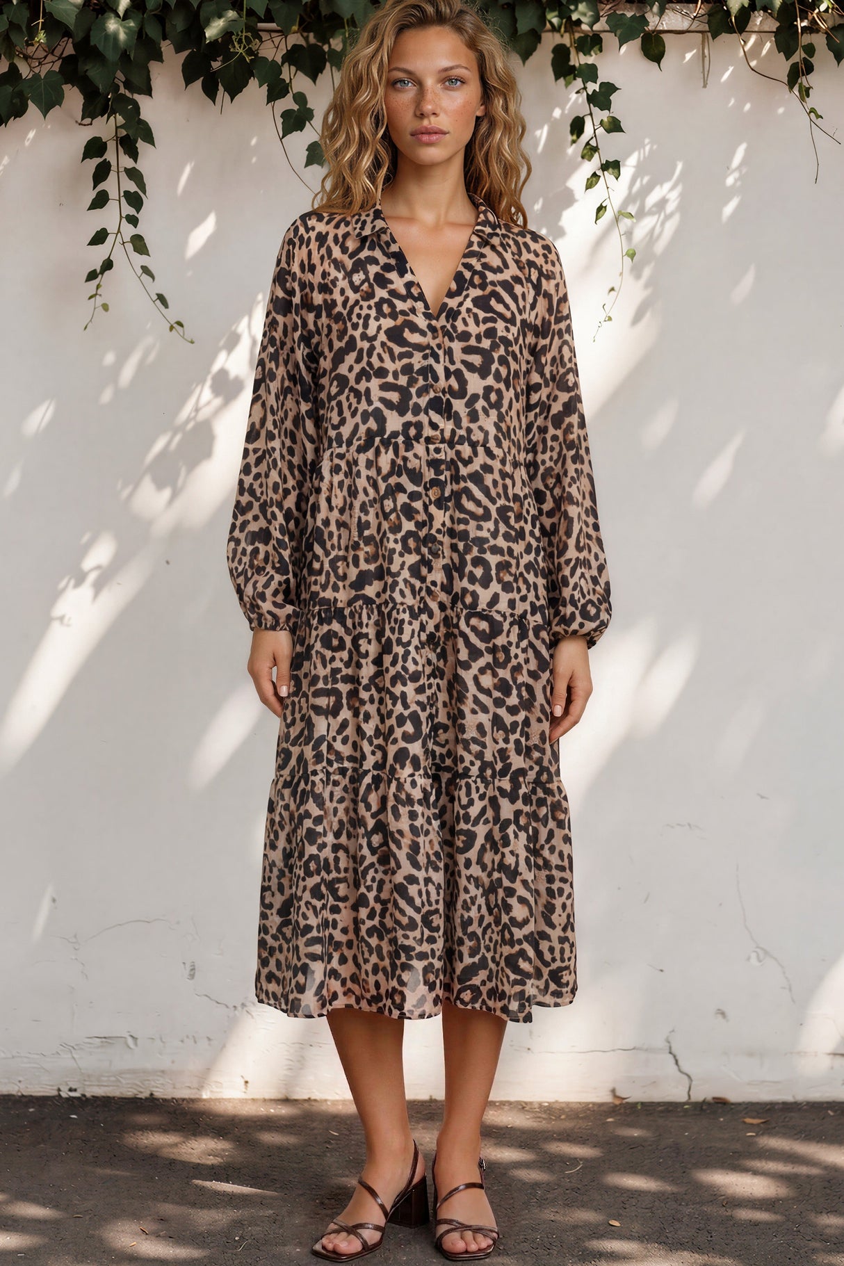 Woman wearing a leopard print dress standing against a white wall with greenery.