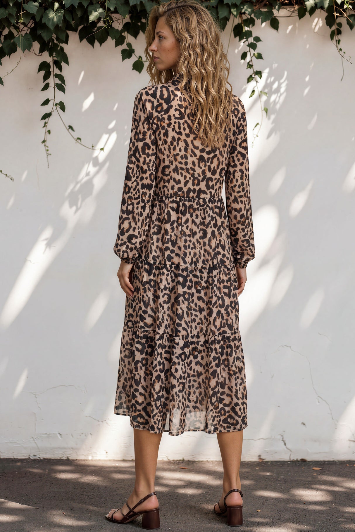 Woman wearing a leopard print dress standing against a white wall with greenery.