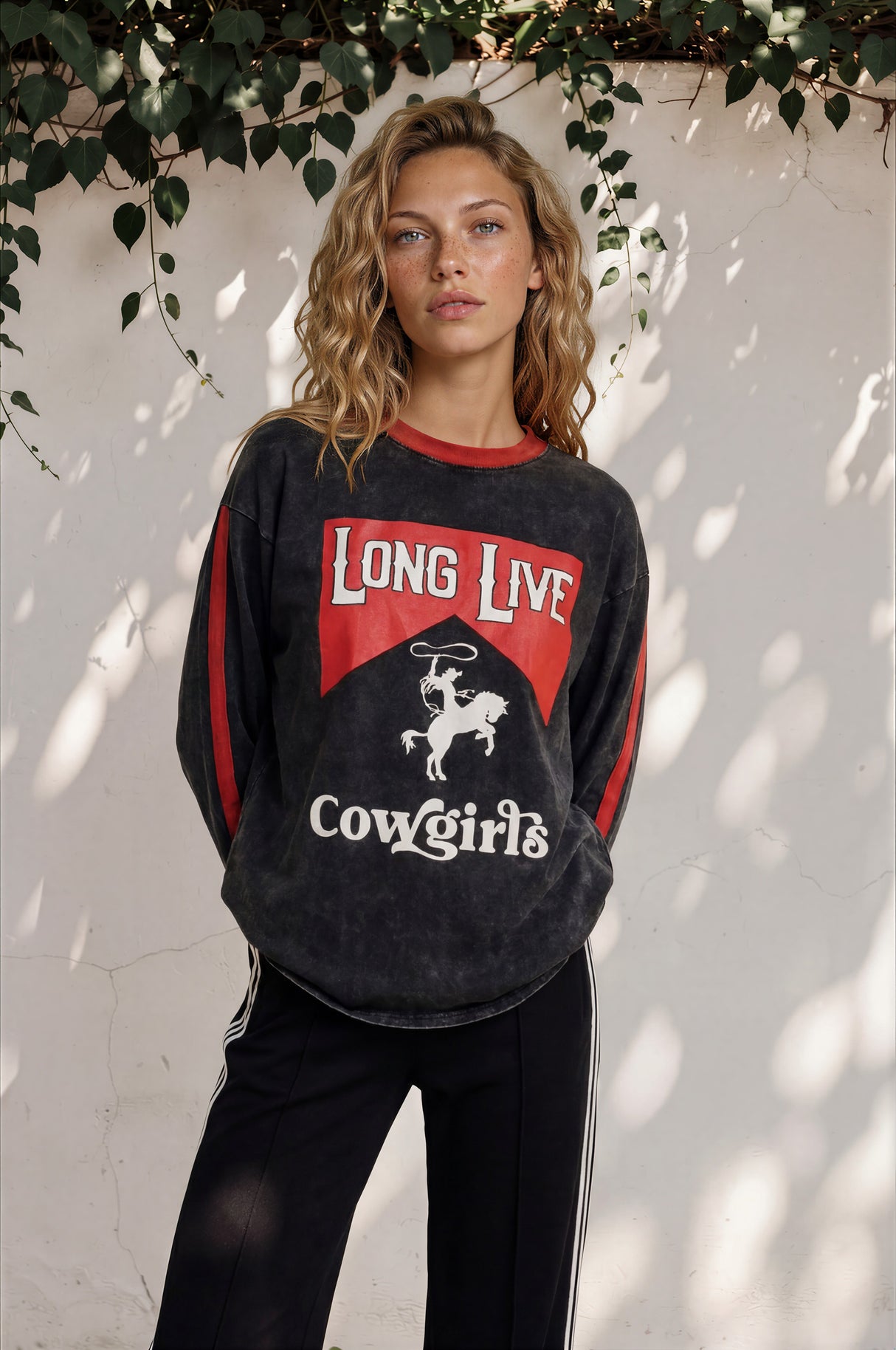 Woman wearing a black sweatshirt with red and white text and graphics, standing against a white wall with greenery.