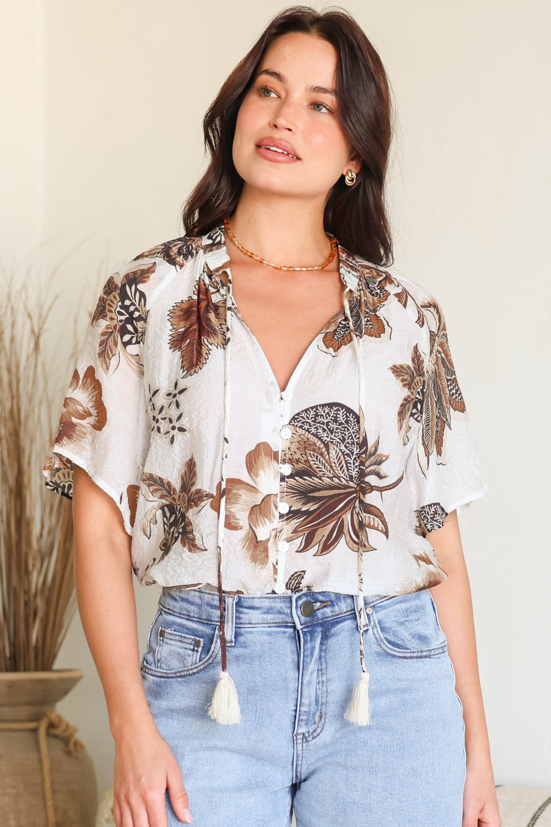 A woman wearing a white short-sleeve button-up blouse with brown and beige botanical leaf patterns and drawstring tassels, paired with light-wash denim shorts, standing in a brightly lit indoor setting.